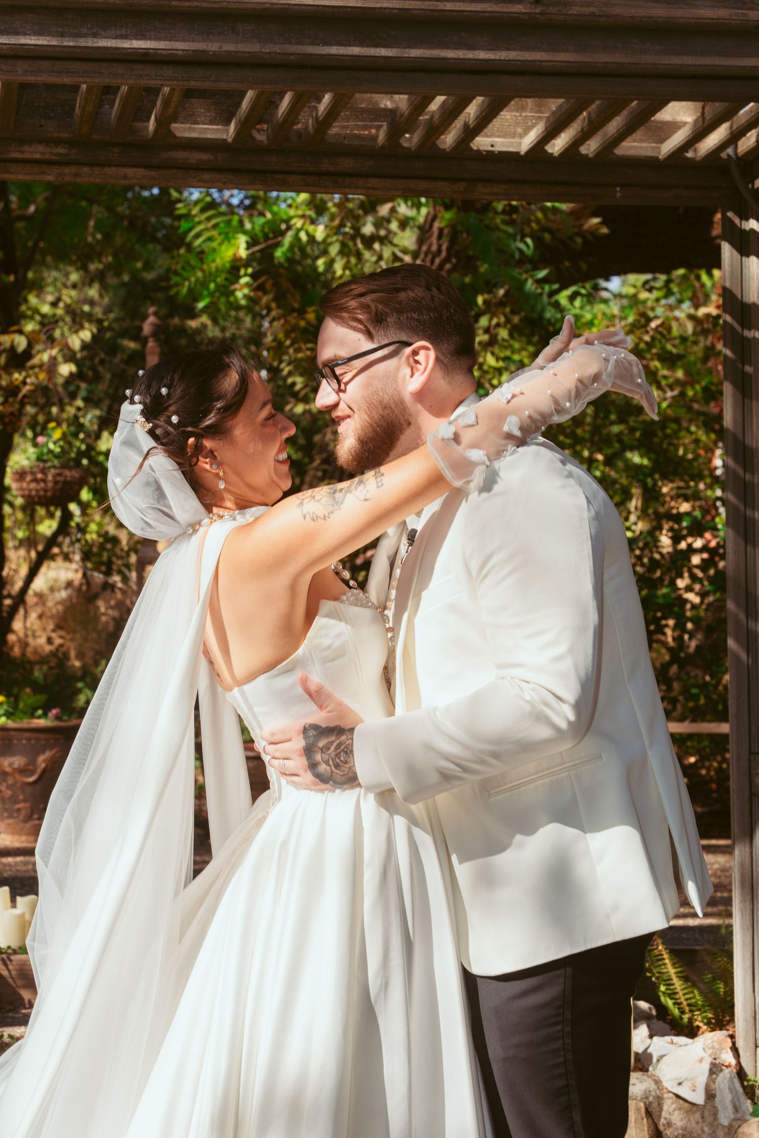 Intimate wedding moment captured under wooden pergola with flowing white dress and formal attire.
