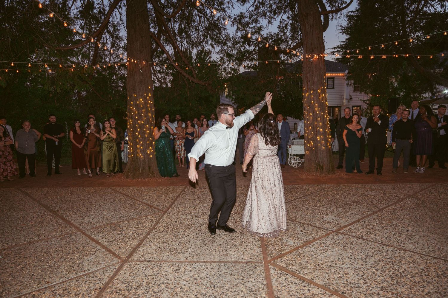 Evening wedding reception with guests dancing under glowing string lights on a patio.