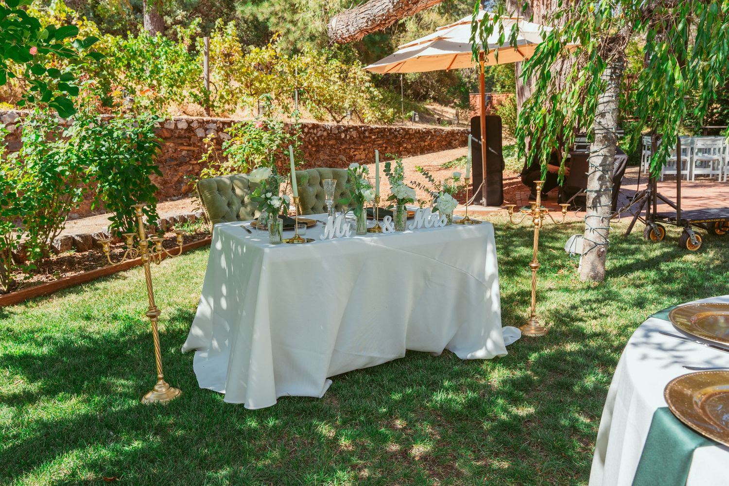 Outdoor garden wedding reception setup with round tables and white tablecloths under trees.
