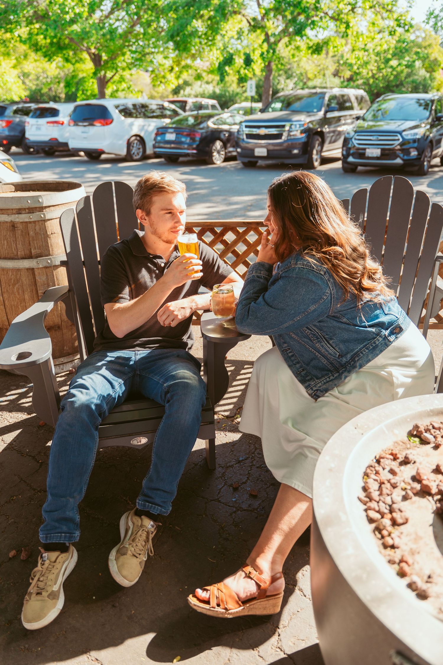 Two people sit casually on wooden benches at an outdoor venue with parked cars in the background.