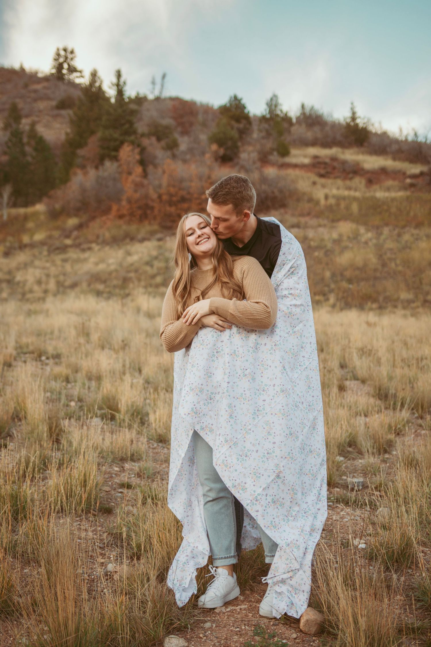 Couple sharing joyful moments while wrapped in white lace blanket in mountain field.