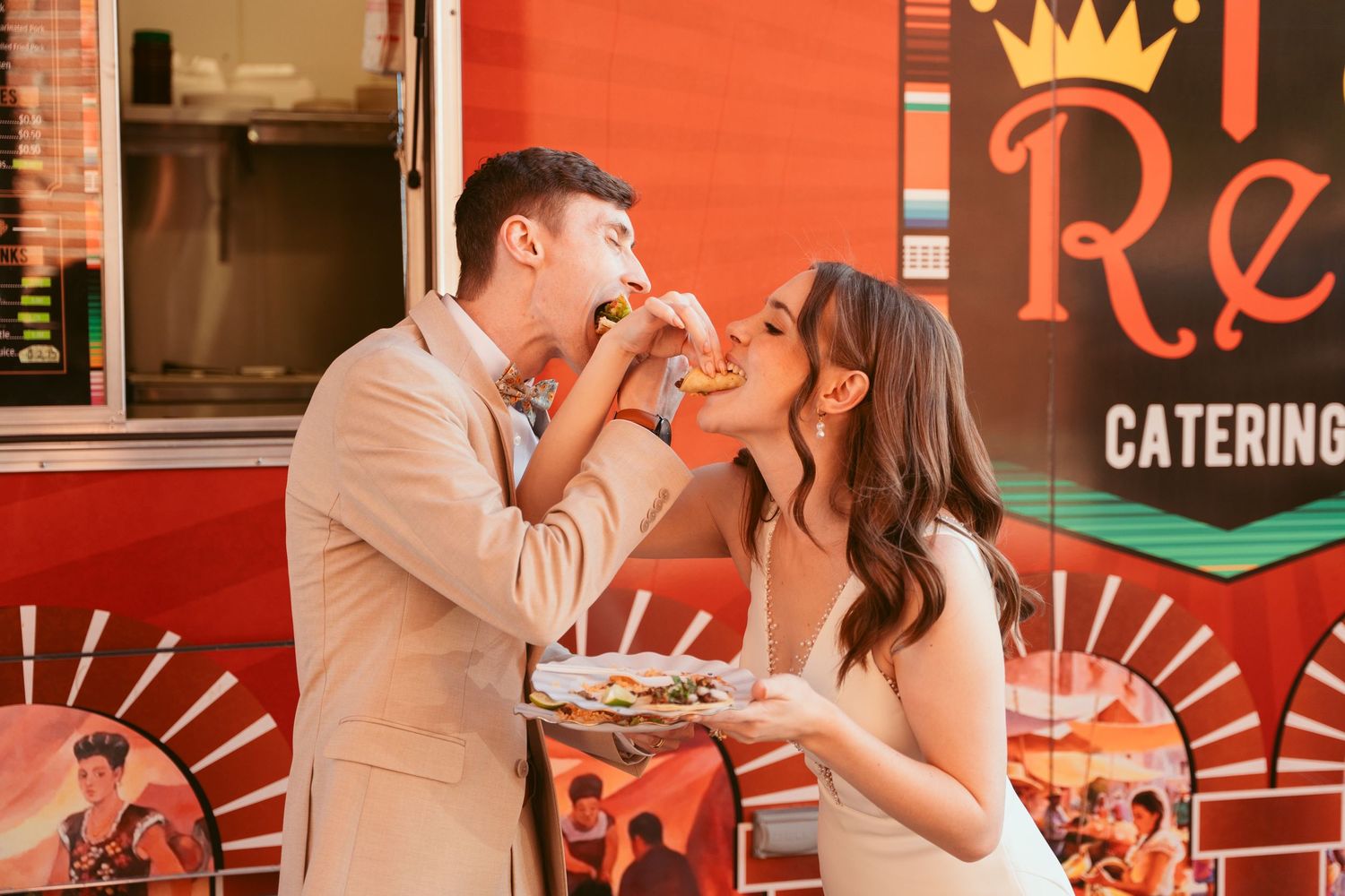 Newlyweds sharing a casual meal from a bright red taco truck during their wedding festivities.