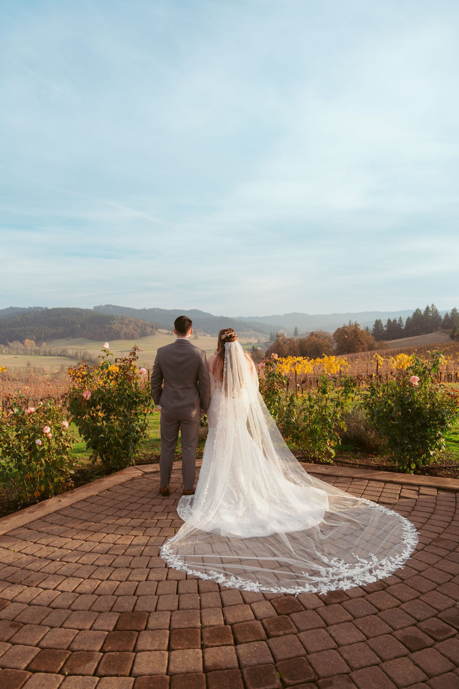 Wedding couple stands on brick patio overlooking scenic vineyard landscape.