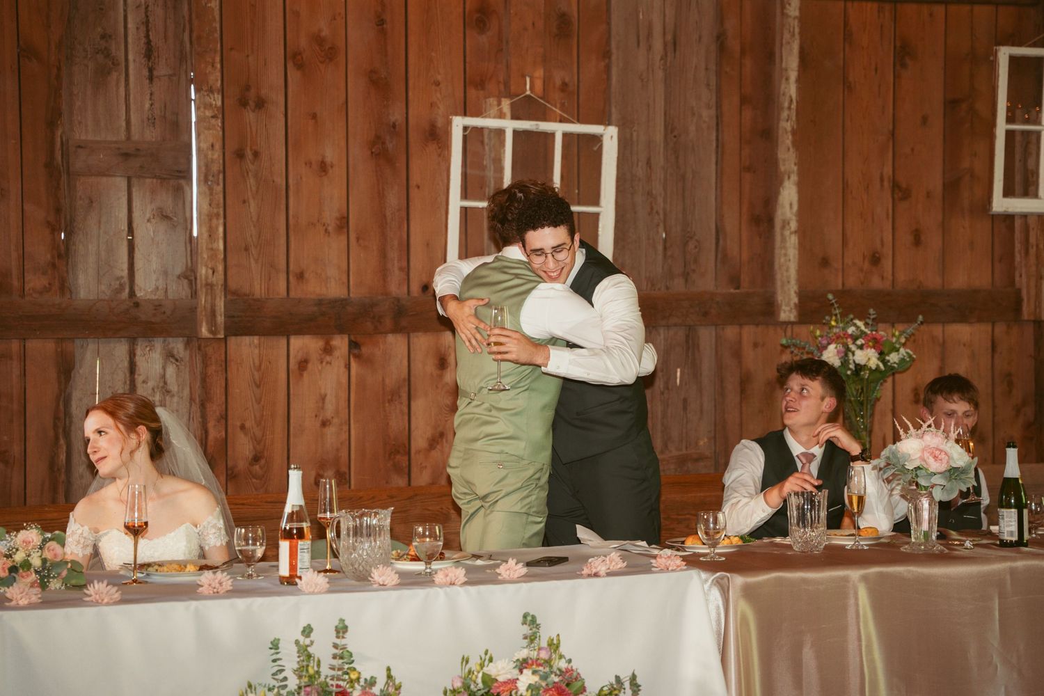 Wedding party members share emotional toasts at rustic barn reception with wooden walls and floral centerpieces.