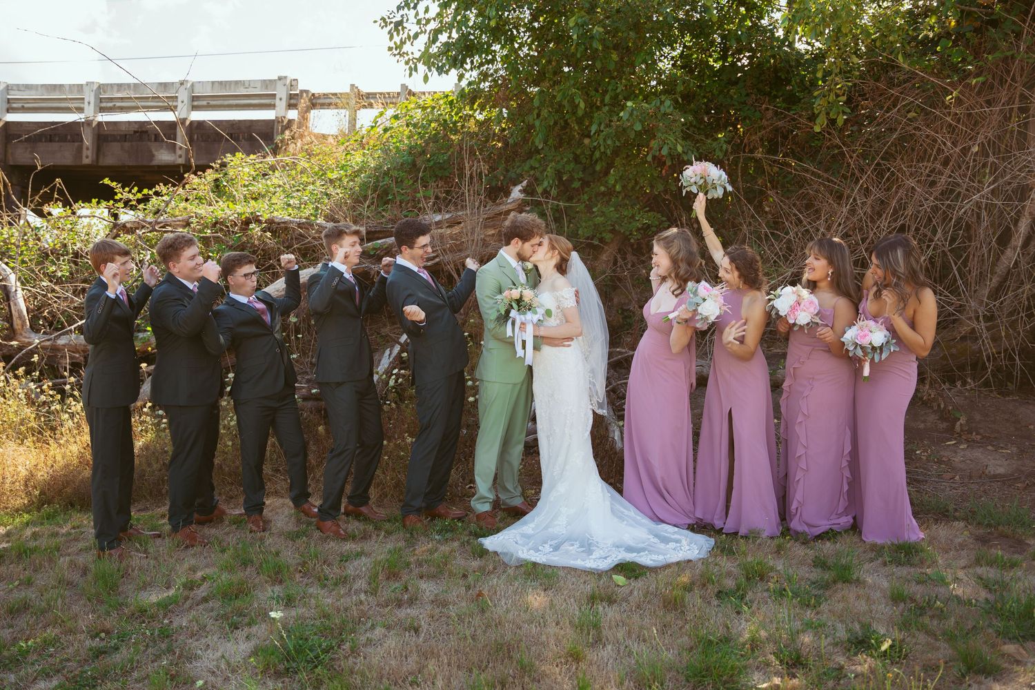 Wedding party stands in line against natural outdoor backdrop with trees and wooden fence.