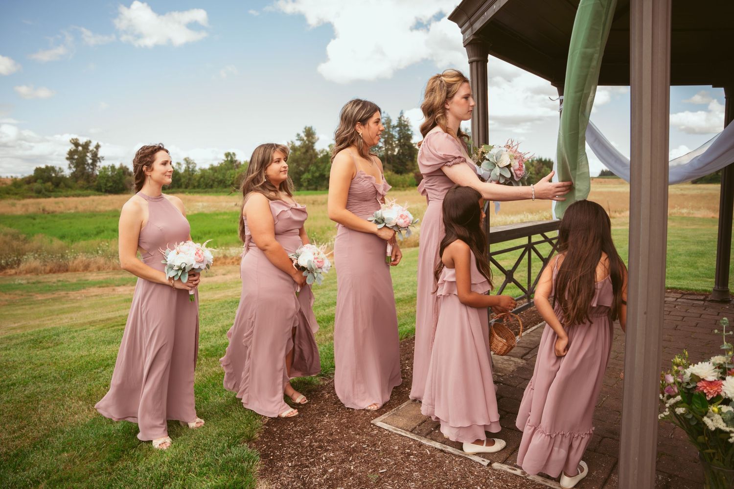Bridesmaids in pink dresses standing in line under wooden gazebo.