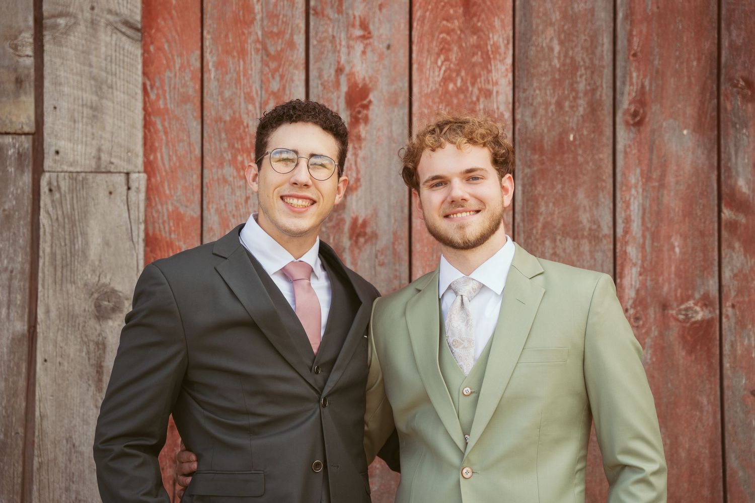 Happy couple in formal attire smile together against weathered red barn background.
