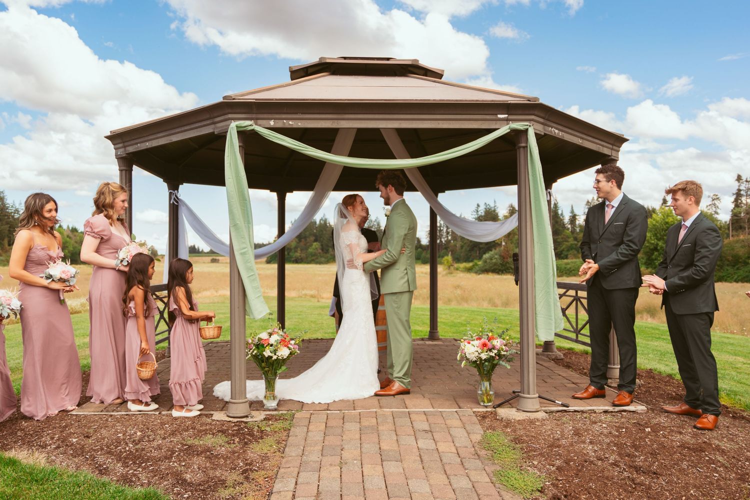 Wedding ceremony at outdoor gazebo with bridal party in pink dresses and groomsmen in grey suits.