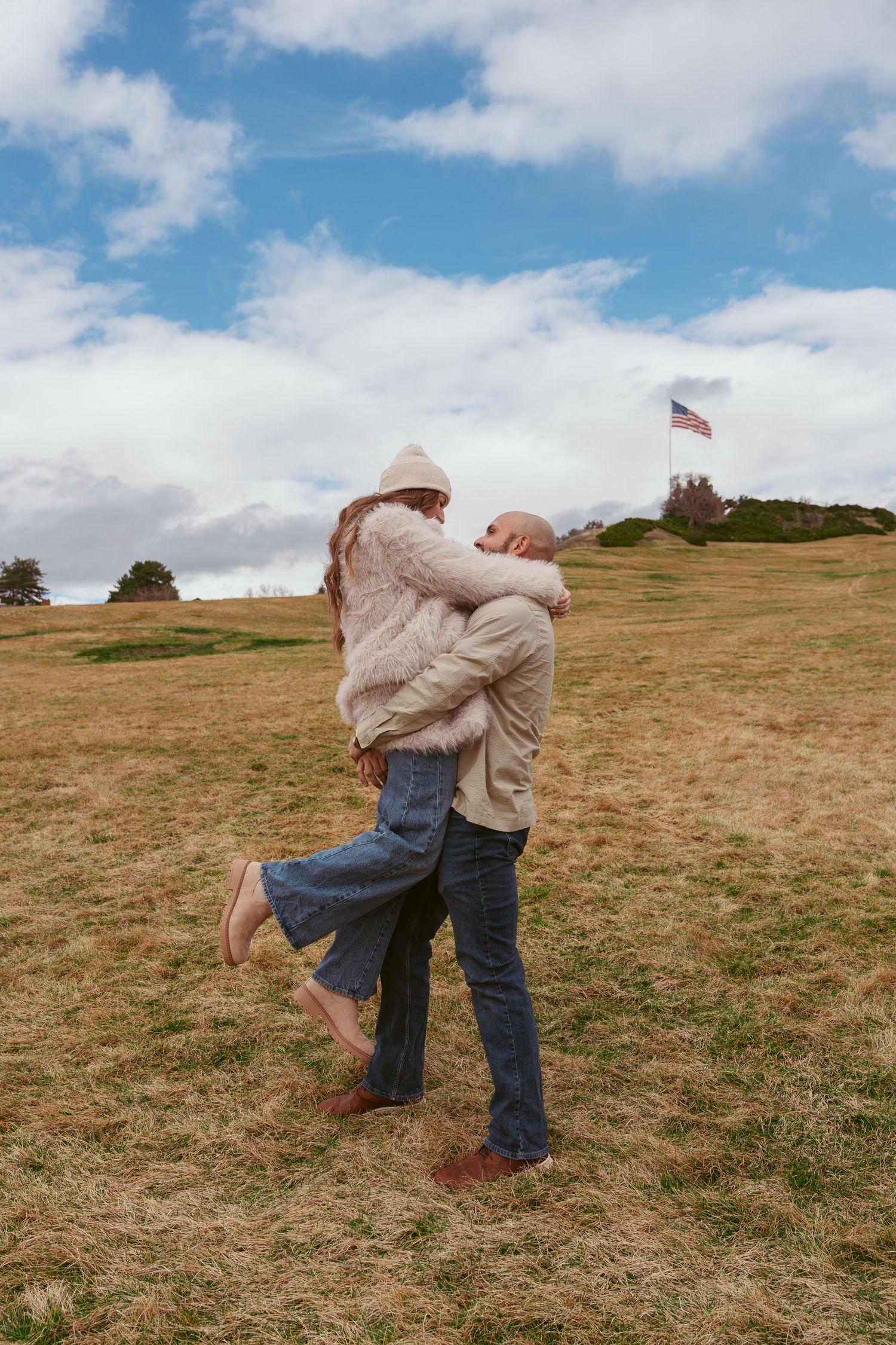 Couple embracing on a grassy hillside under blue skies and white clouds.