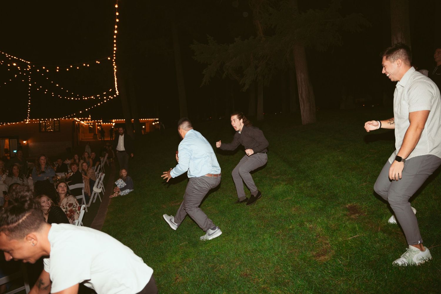 Wedding guests dancing energetically on a dark dance floor with string lights overhead.
