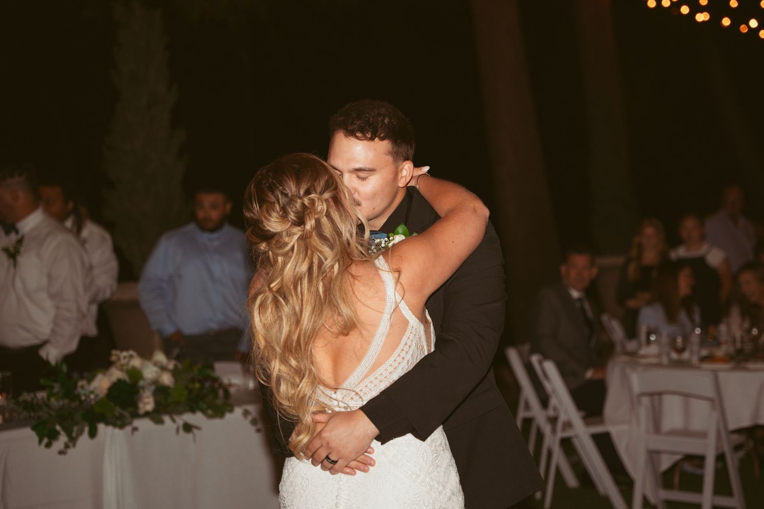 A couple shares a romantic dance at a wedding reception under warm lighting with guests seated in the background.