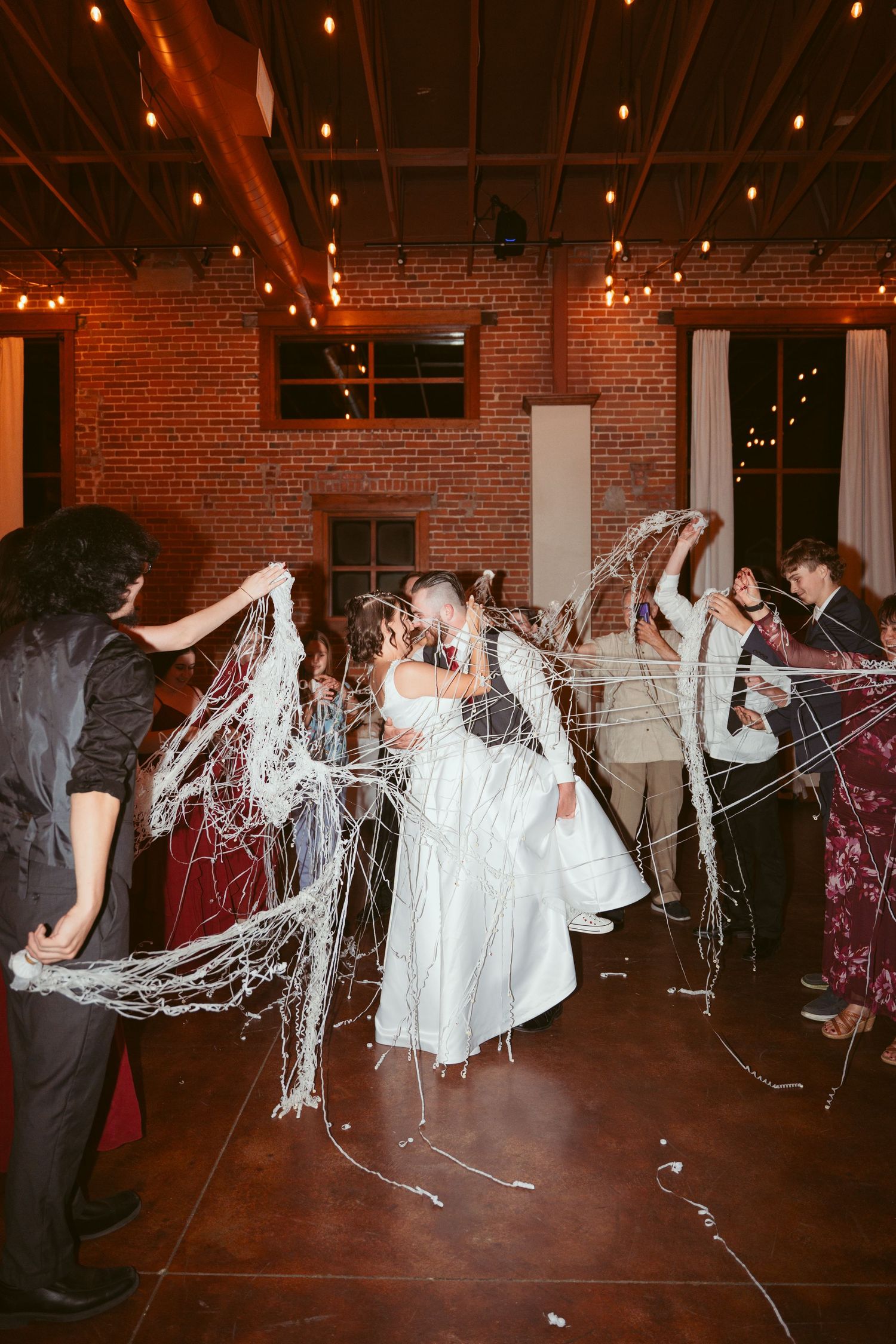 Wedding guests celebrate by throwing white streamers across dance floor in rustic brick venue.