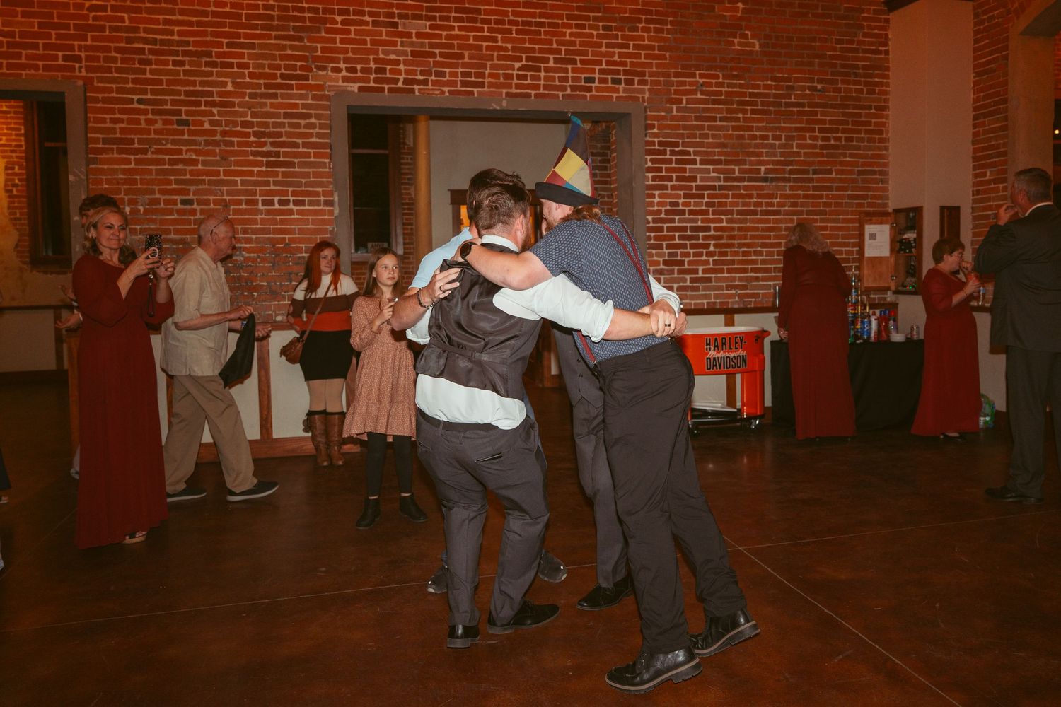Wedding guests dancing together in a rustic brick-walled venue with string lights overhead.