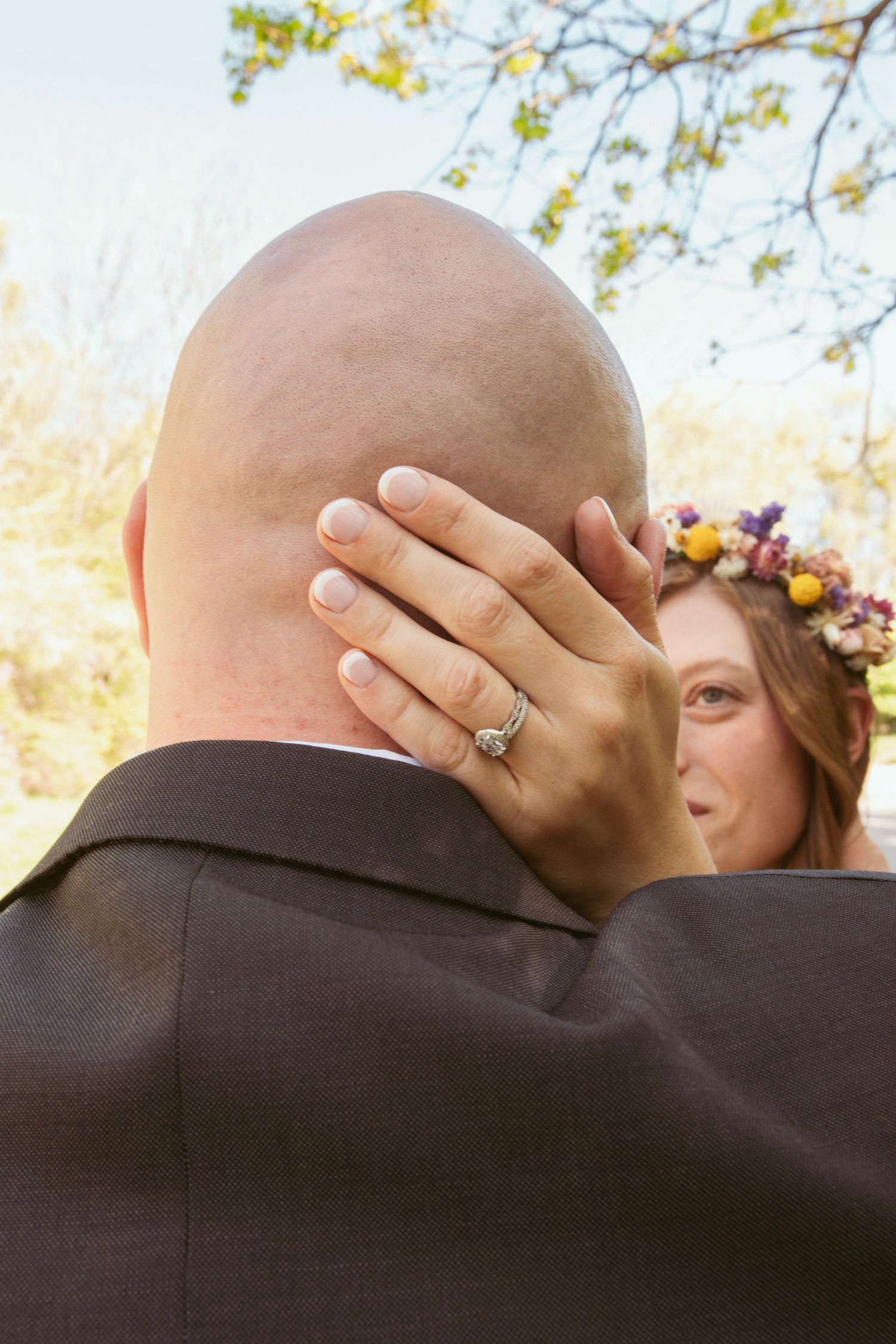 A bride wearing a floral crown playfully covers her groom's face while showing off her engagement ring.