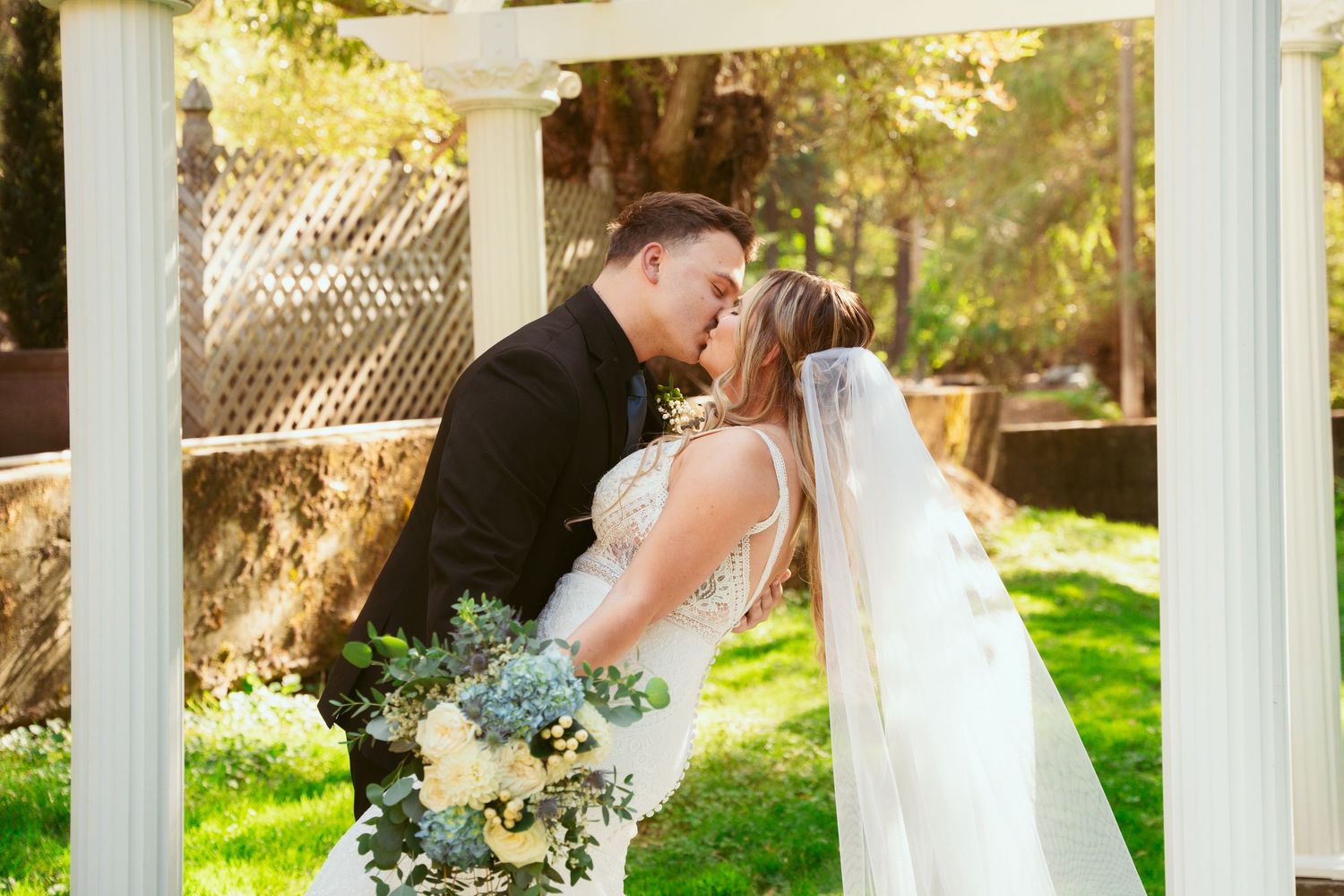 A romantic wedding couple shares a tender kiss under white columns in a sunlit garden setting.