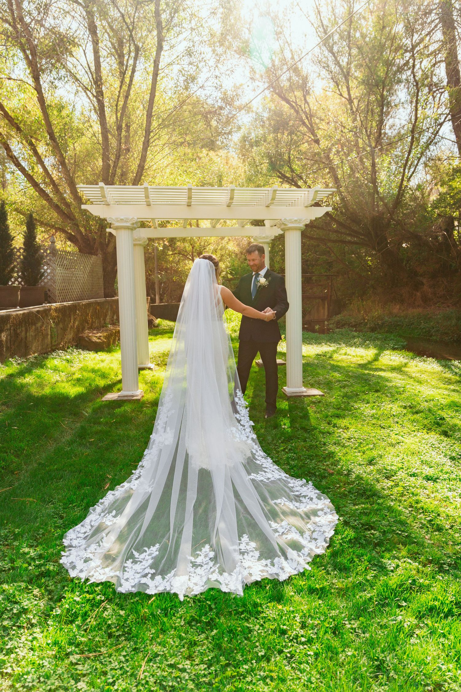 A flowing cathedral length lace veil trails behind a bride in a garden setting with white pergola and green grass.