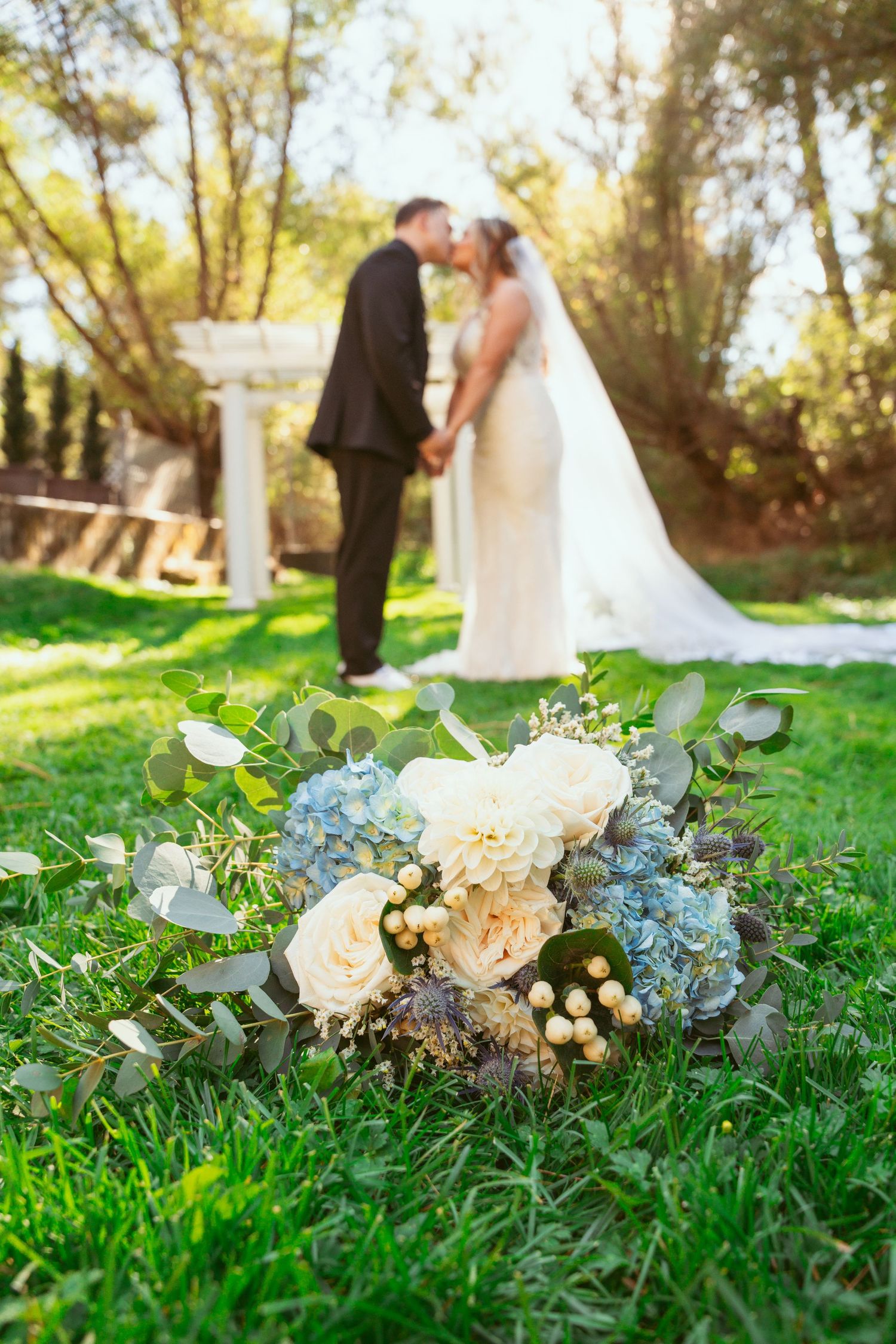 Wedding bouquet with white roses and eucalyptus lies on grass with newlyweds in soft focus background.