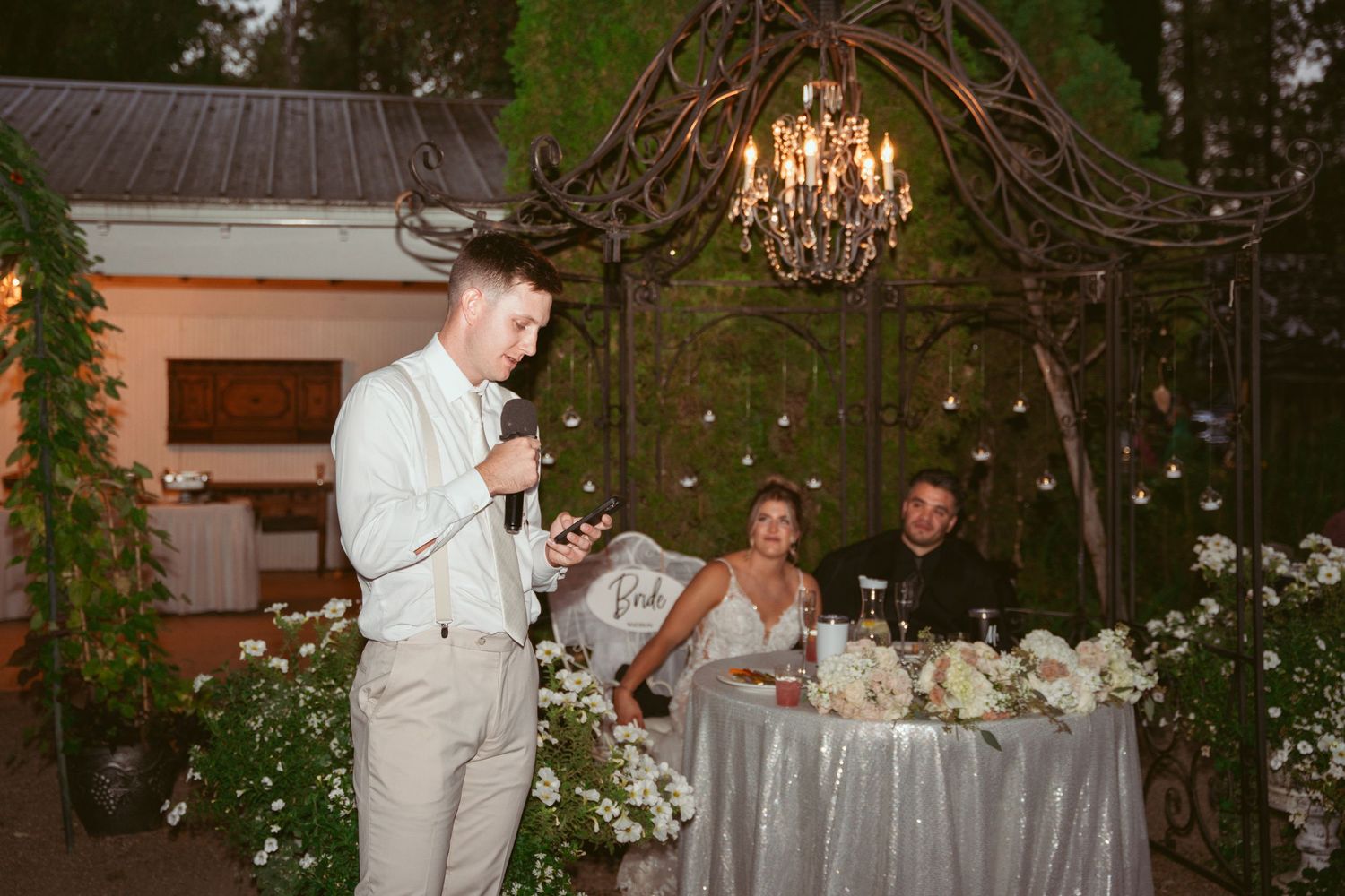 A wedding reception speech being given under a rustic wooden arch with twinkling lights and chandeliers.