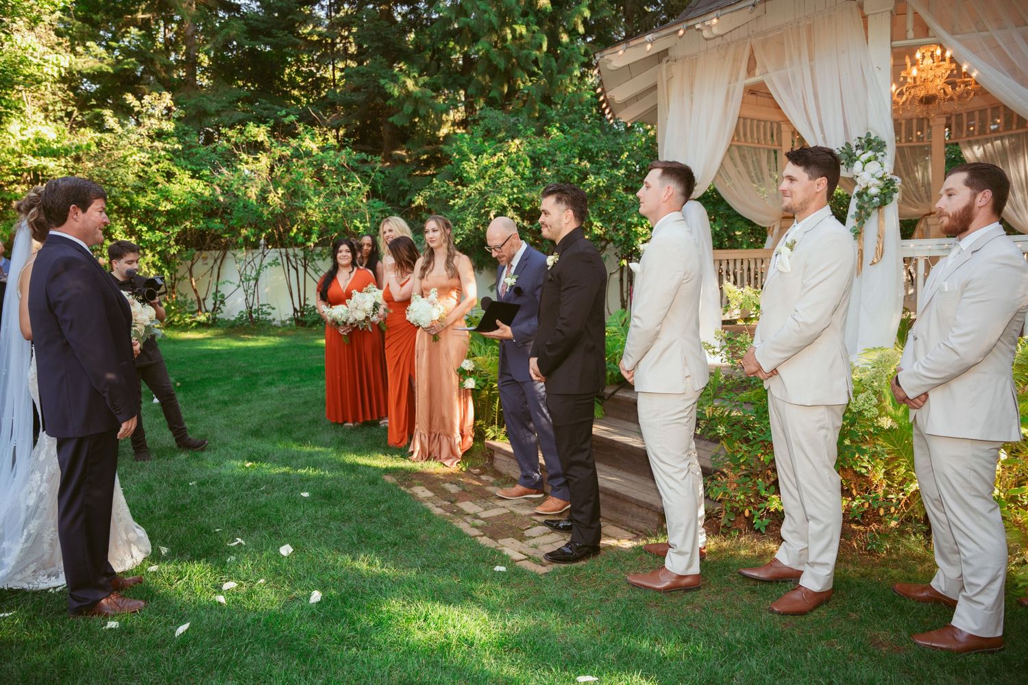 Wedding party stands in formation during outdoor ceremony with bridesmaids in rust dresses and groomsmen in light suits.