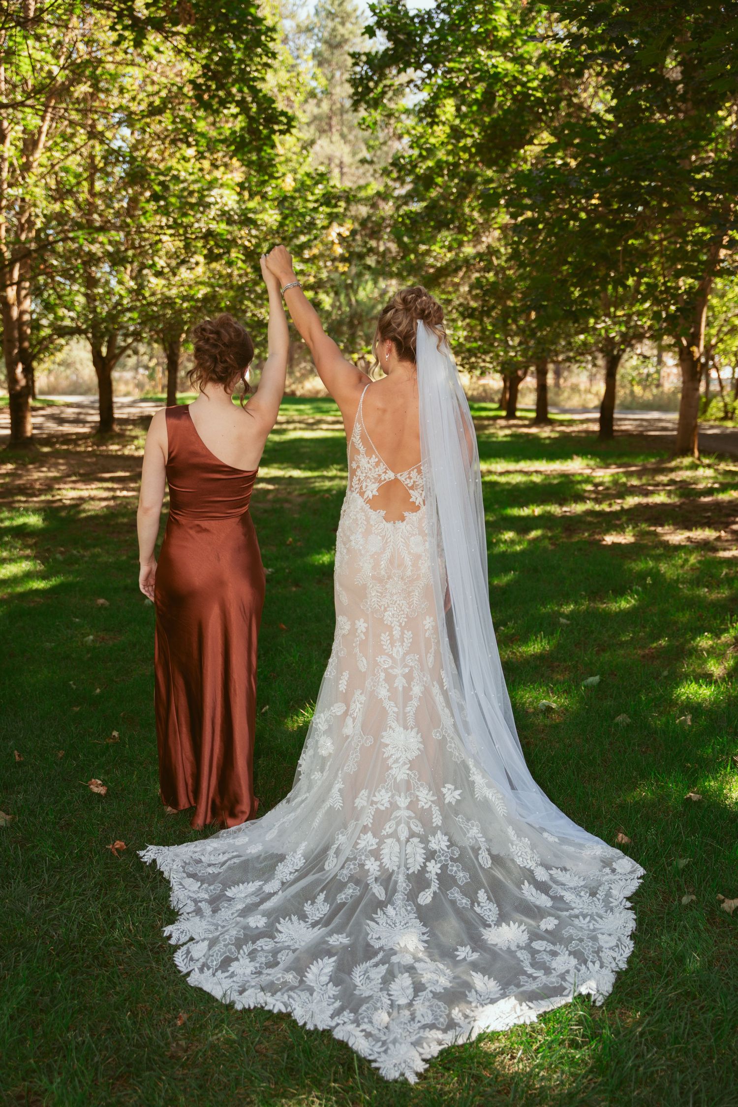 Two people in wedding attire stand together in a sunlit orchard, showcasing the intricate lace details of a flowing white gown.