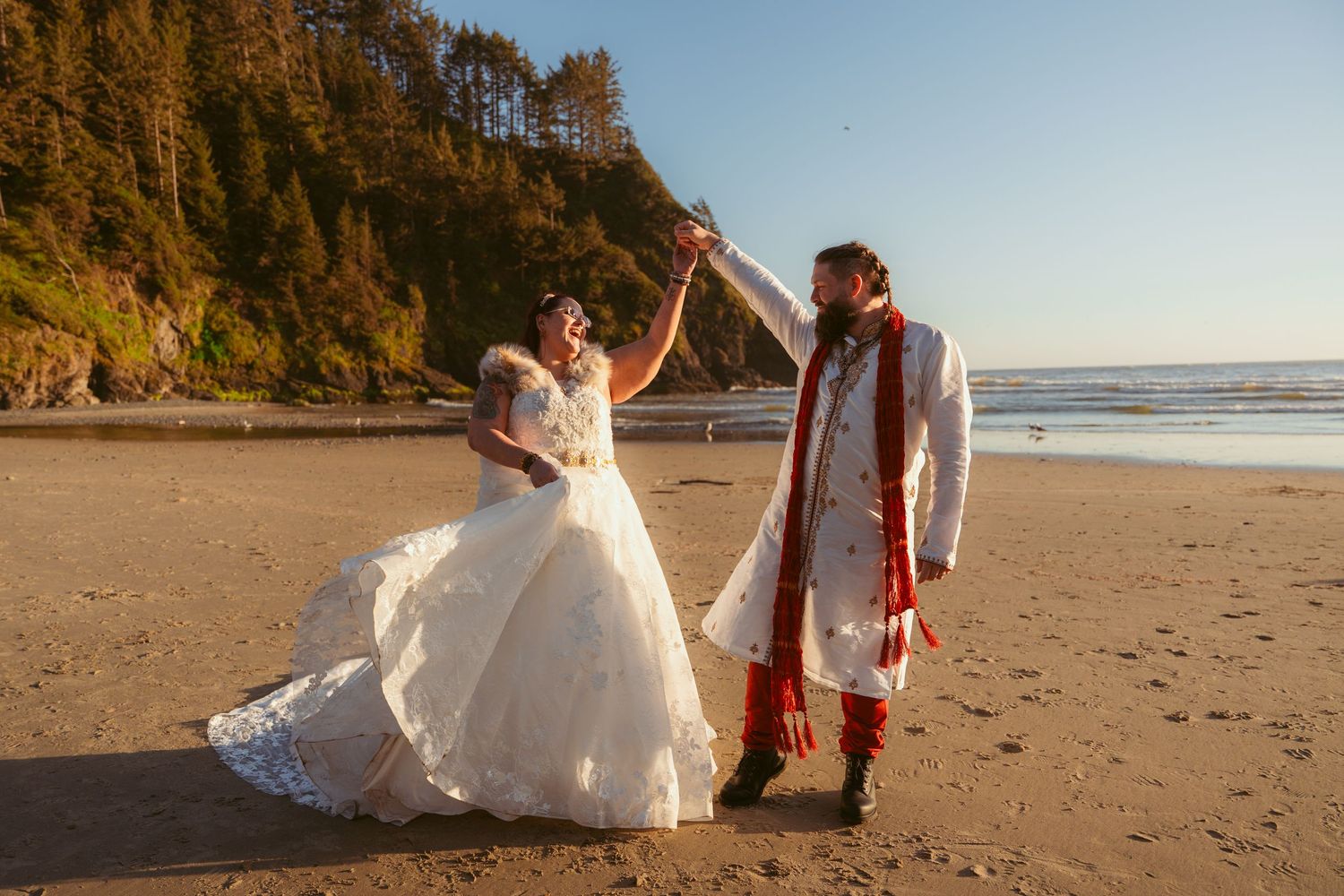 Two people share a joyful moment on a sandy beach with cliffs and ocean waves in the background.