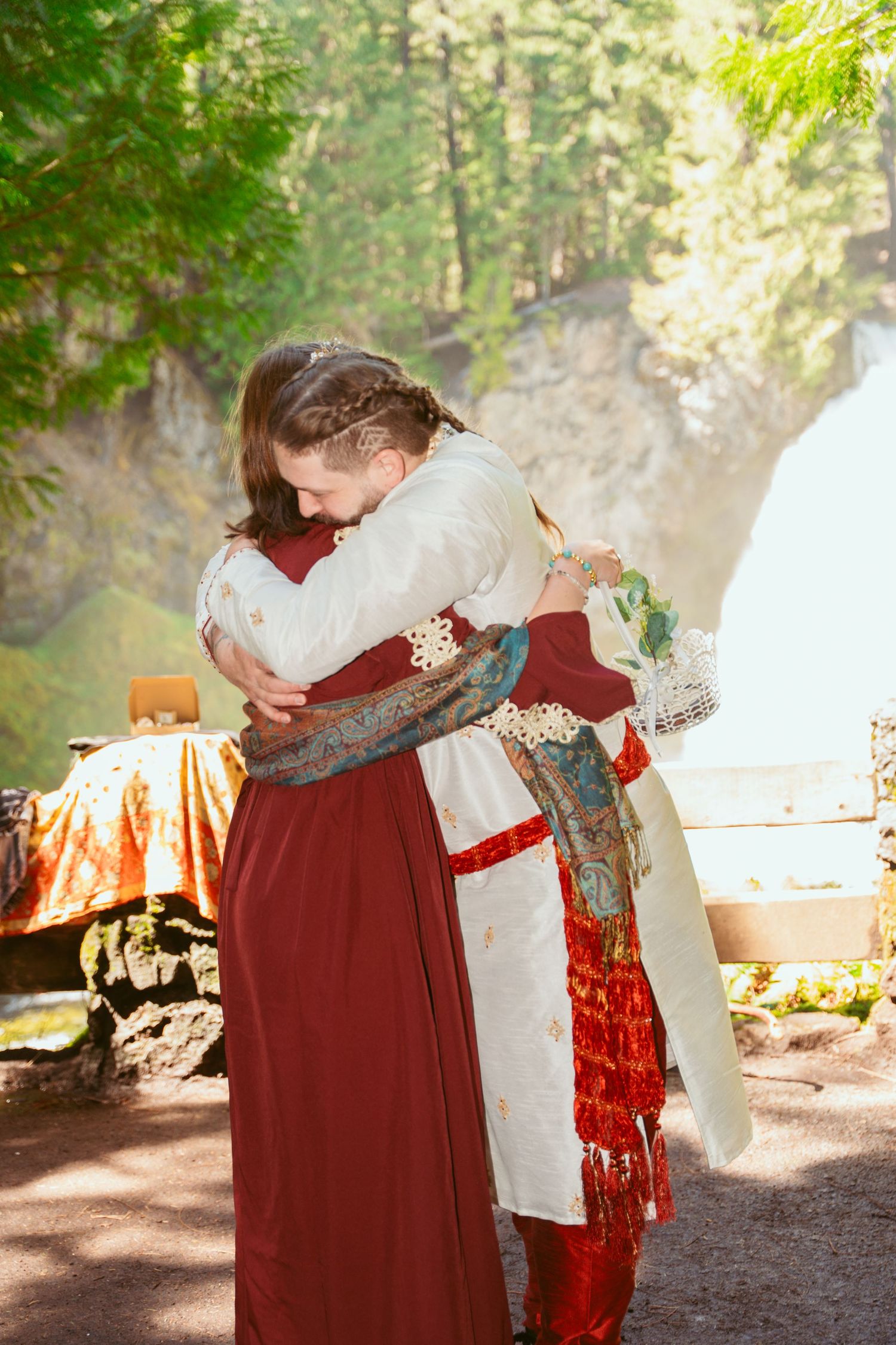 Wedding party members in burgundy and white medieval attire embrace during outdoor celebrations.