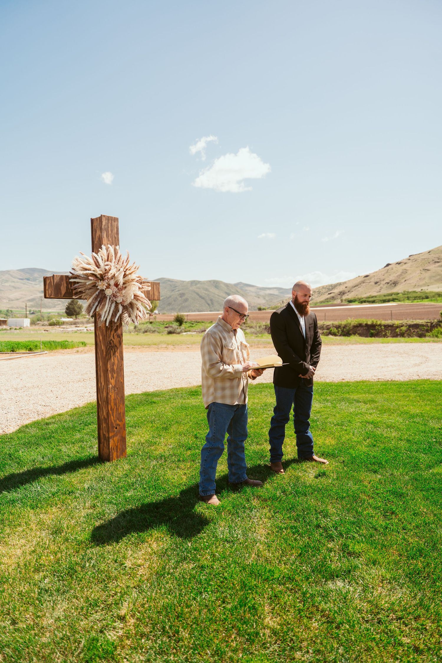 Rustic wooden cross stands in green grass field during outdoor wedding ceremony with mountain views.