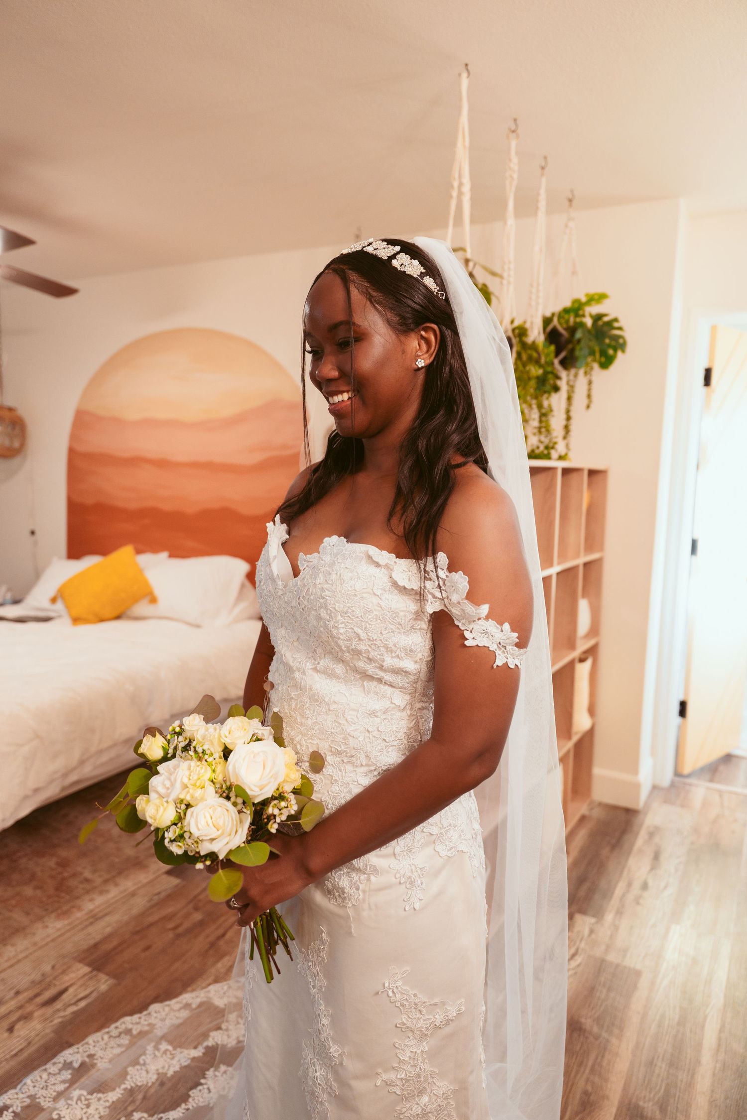 A bride in a white lace dress holds a bouquet in an indoor setting with peach-colored walls.