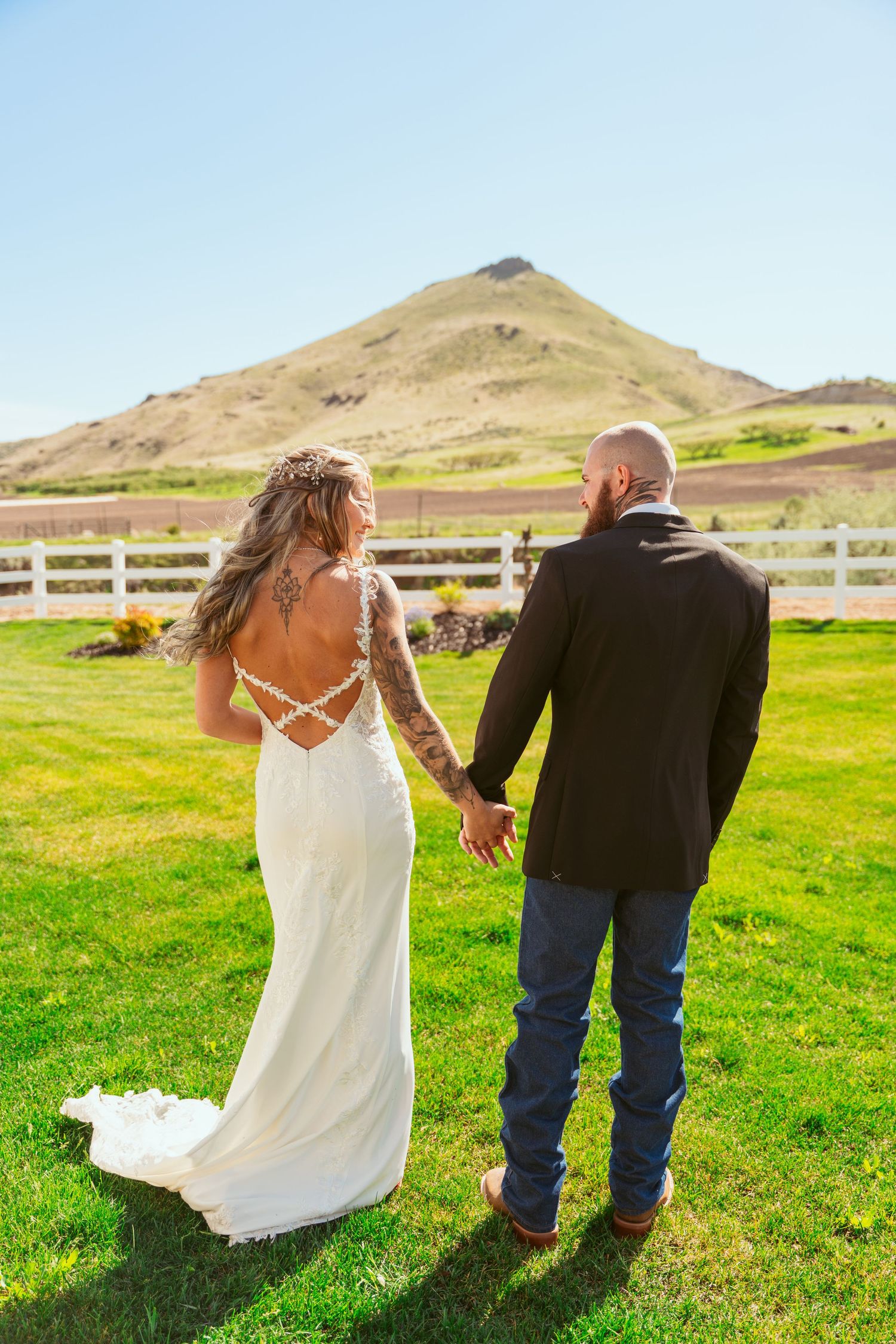 Couple walks together on grass with mountain landscape and white fence in background.
