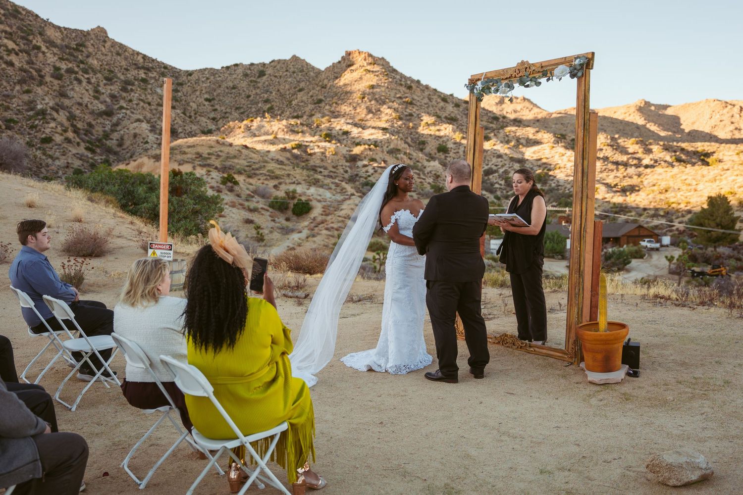 Desert wedding ceremony with guests watching the couple exchange vows under a wooden arch frame.