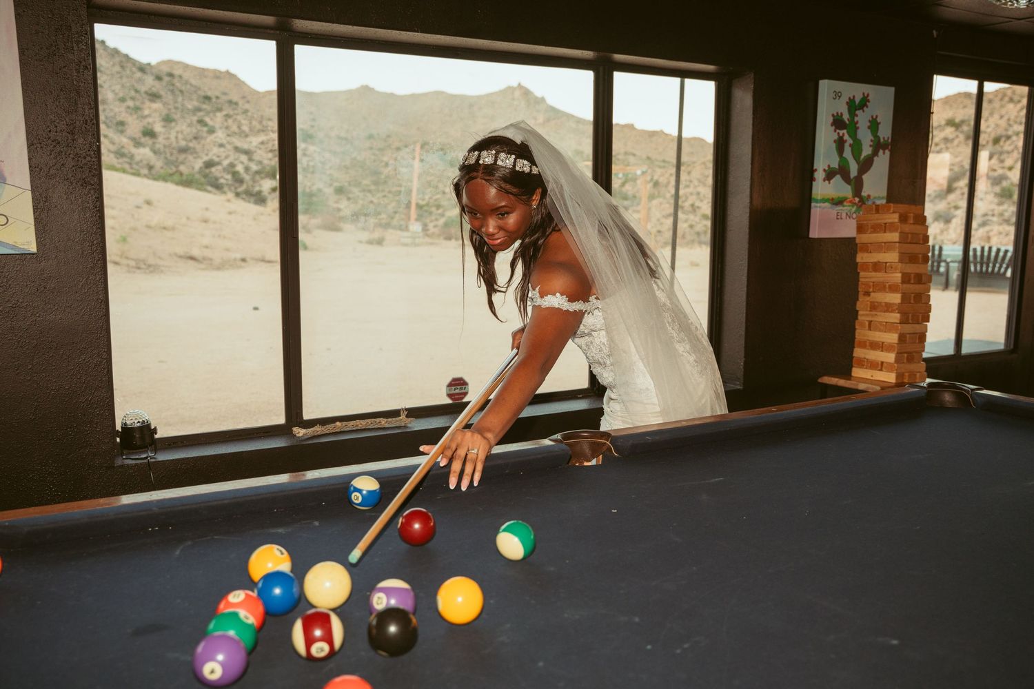 A bride in white dress plays pool while wearing her wedding veil.