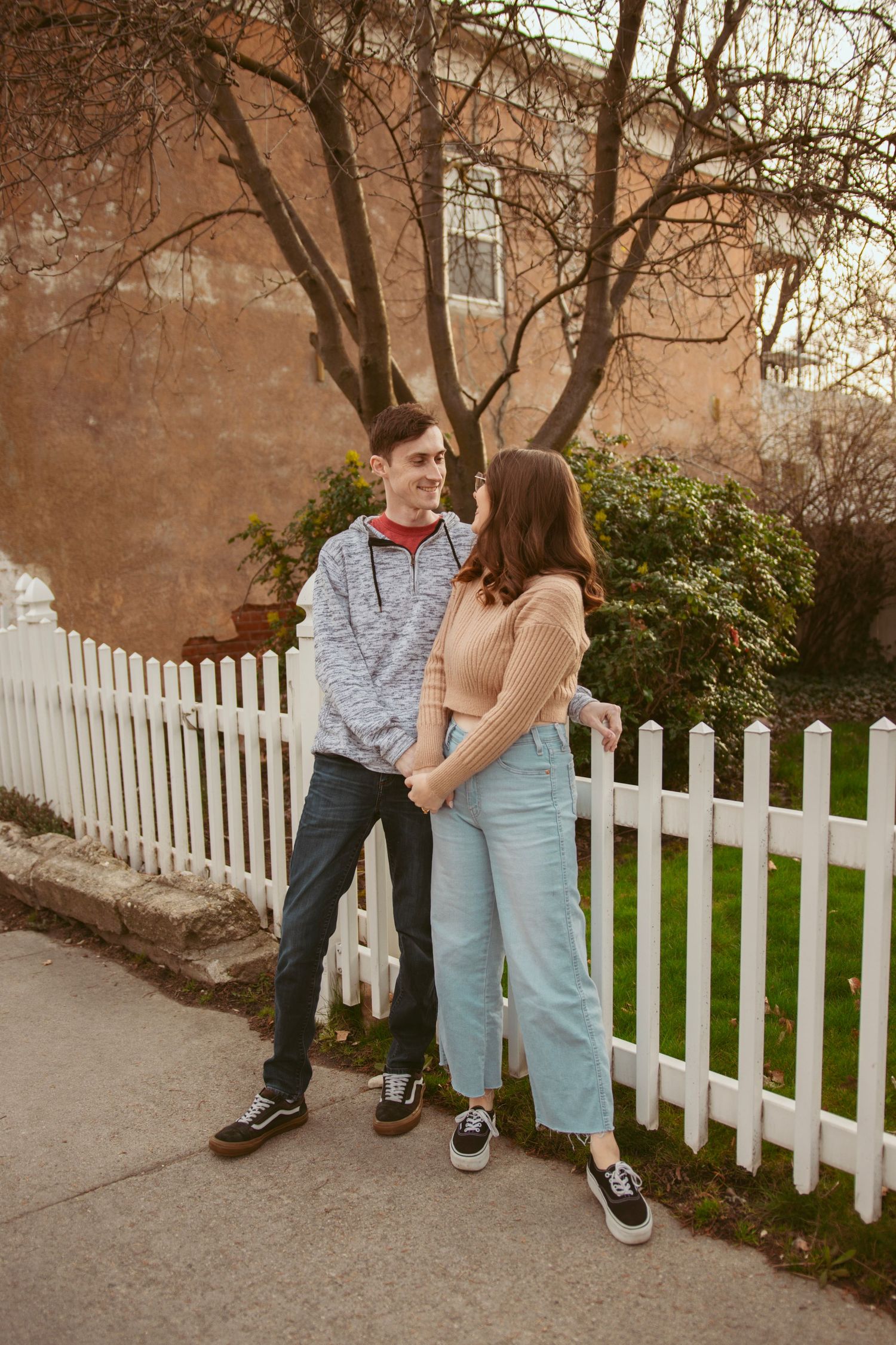 A couple in casual attire poses together by a white picket fence on a sunny day.