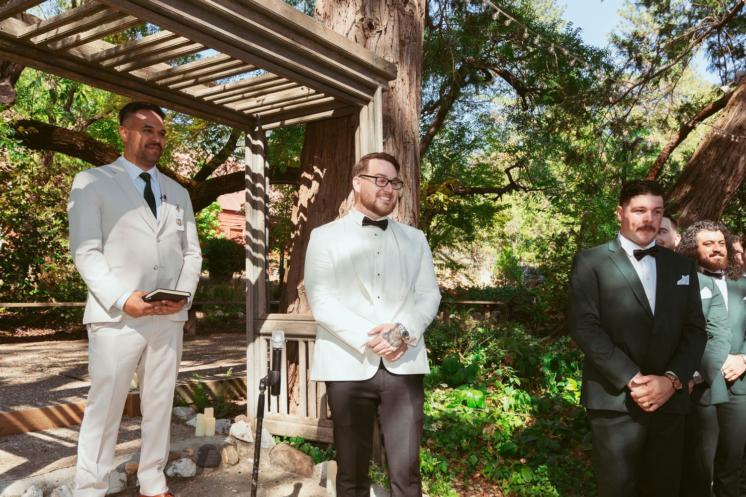 A group of people in formal attire gather under a rustic wooden pergola during an outdoor celebration.