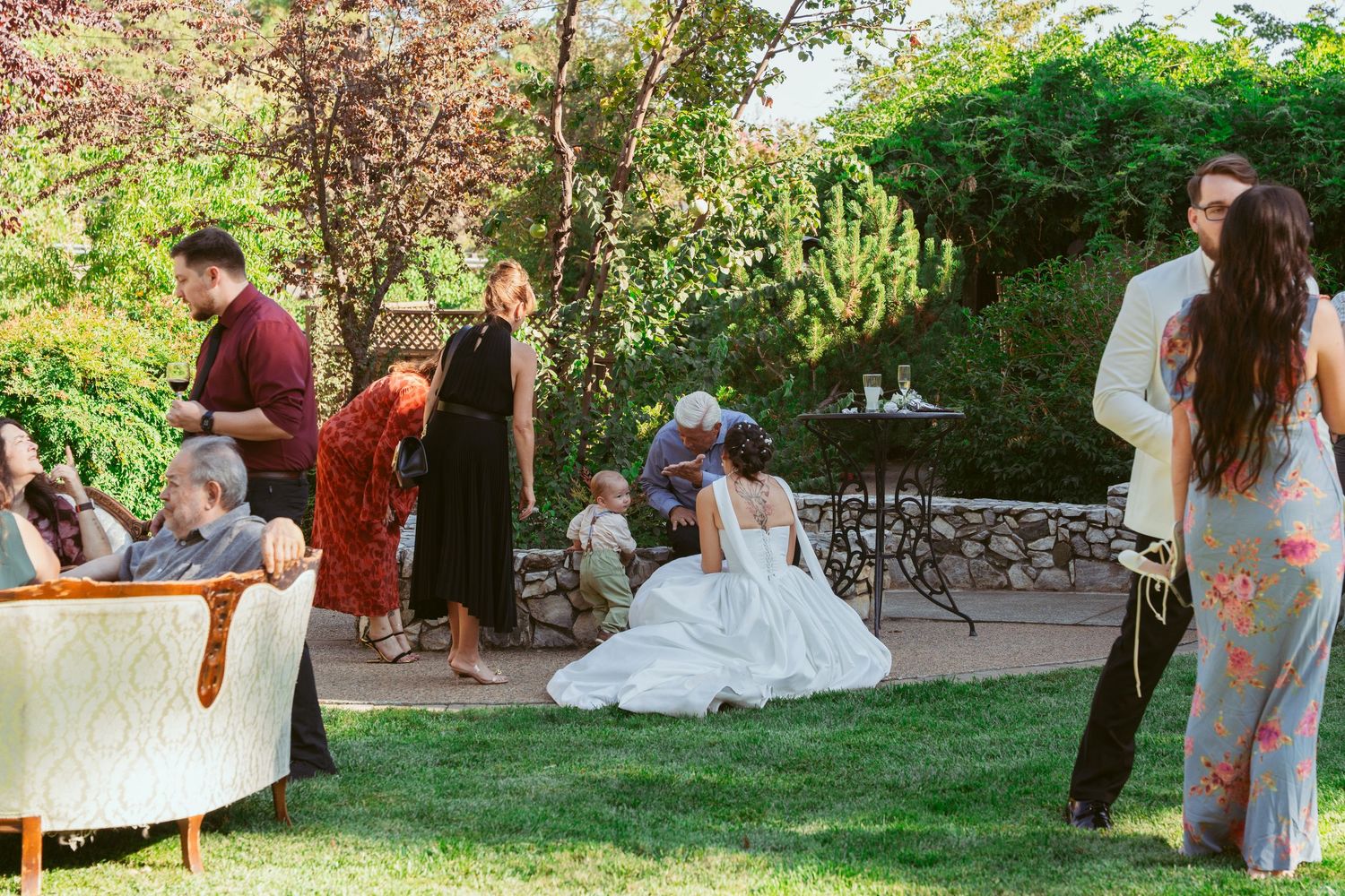 Wedding guests relax and socialize on a lush garden patio during an outdoor reception celebration.