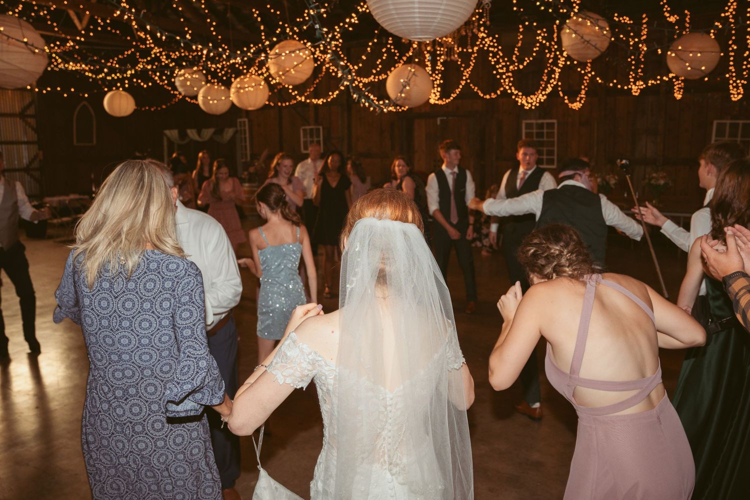 Guests dancing under warm twinkle lights and white paper lanterns at an indoor celebration.