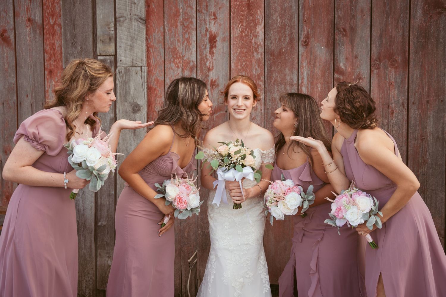 Bridal party in mauve dresses shares laughs while posing with bouquets by wooden wall.