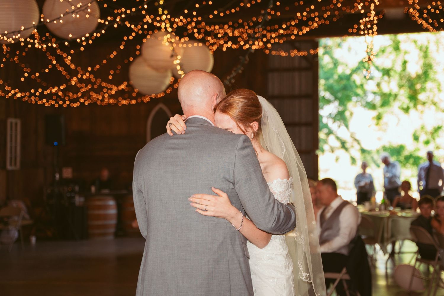 Newlyweds embrace during their wedding dance under glowing string lights in a rustic barn setting.