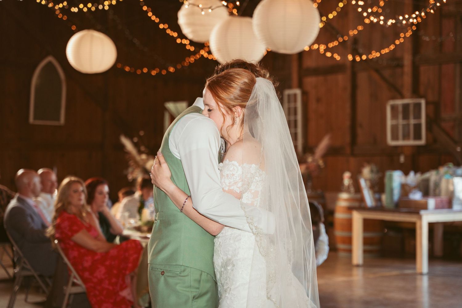 Couple dances together in rustic barn venue decorated with string lights and paper lanterns.