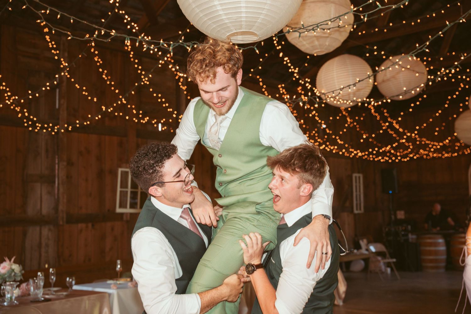Wedding reception guests dance under twinkling string lights and paper lanterns in a rustic barn venue.