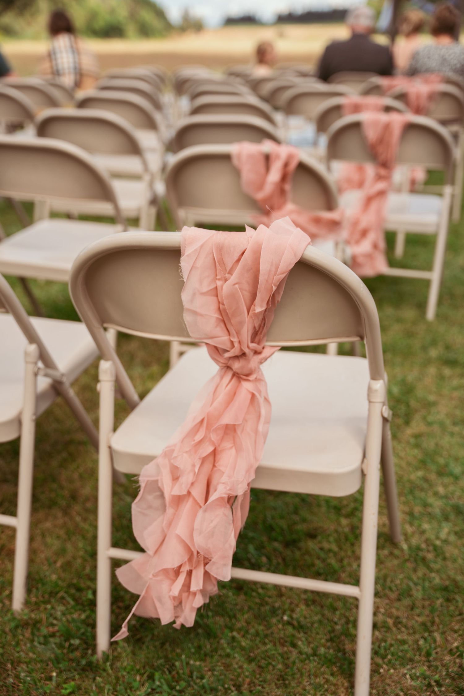 White folding chairs decorated with blush pink fabric bows arranged outdoors on grass.