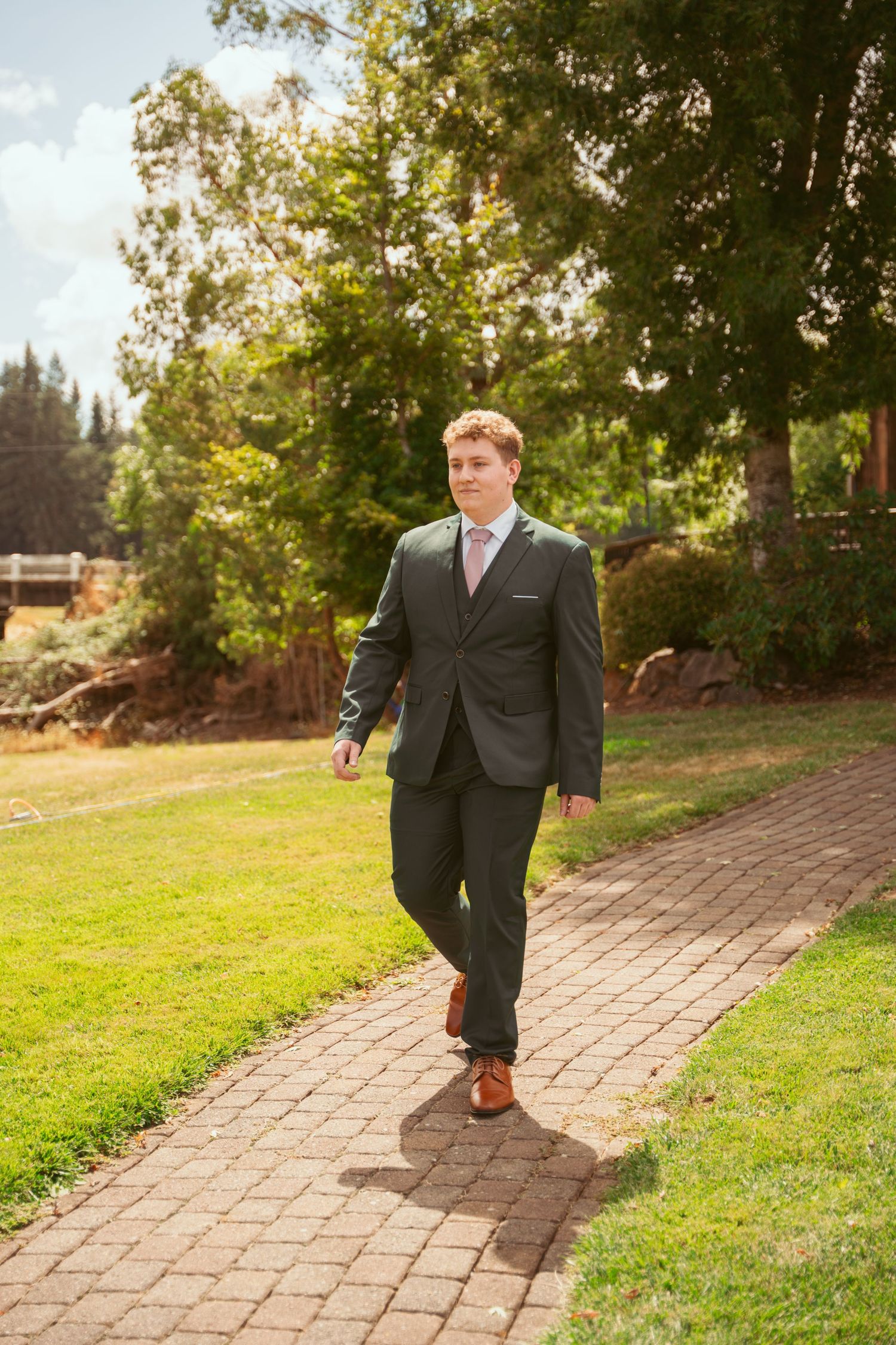 Person in charcoal suit and brown shoes walks on brick path through sunny garden setting.