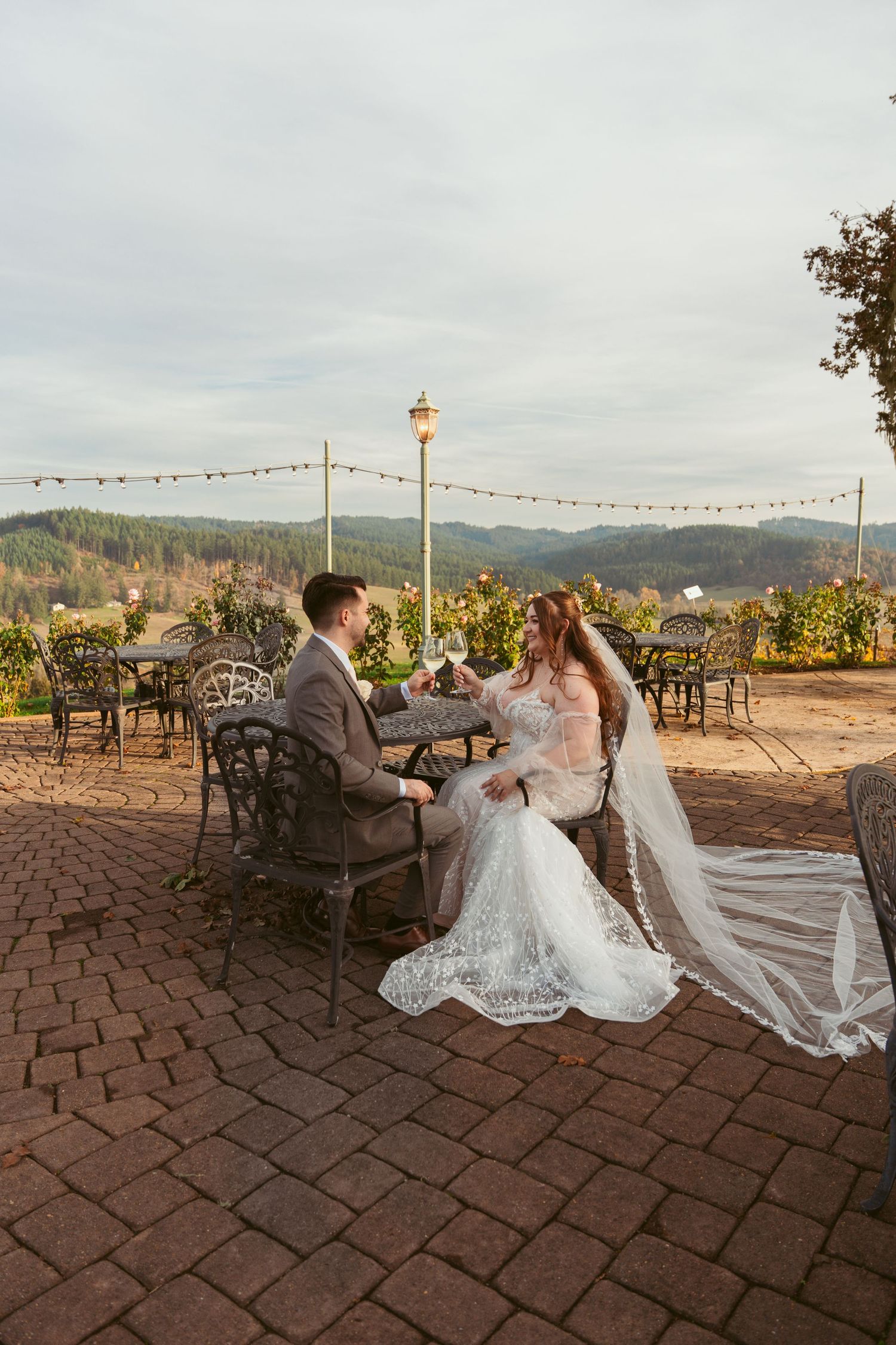 Wedding couple seated at outdoor vineyard patio with string lights overhead.