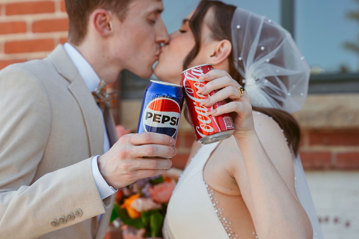 A playful wedding photo featuring Pepsi and Coca-Cola cans during a romantic moment.