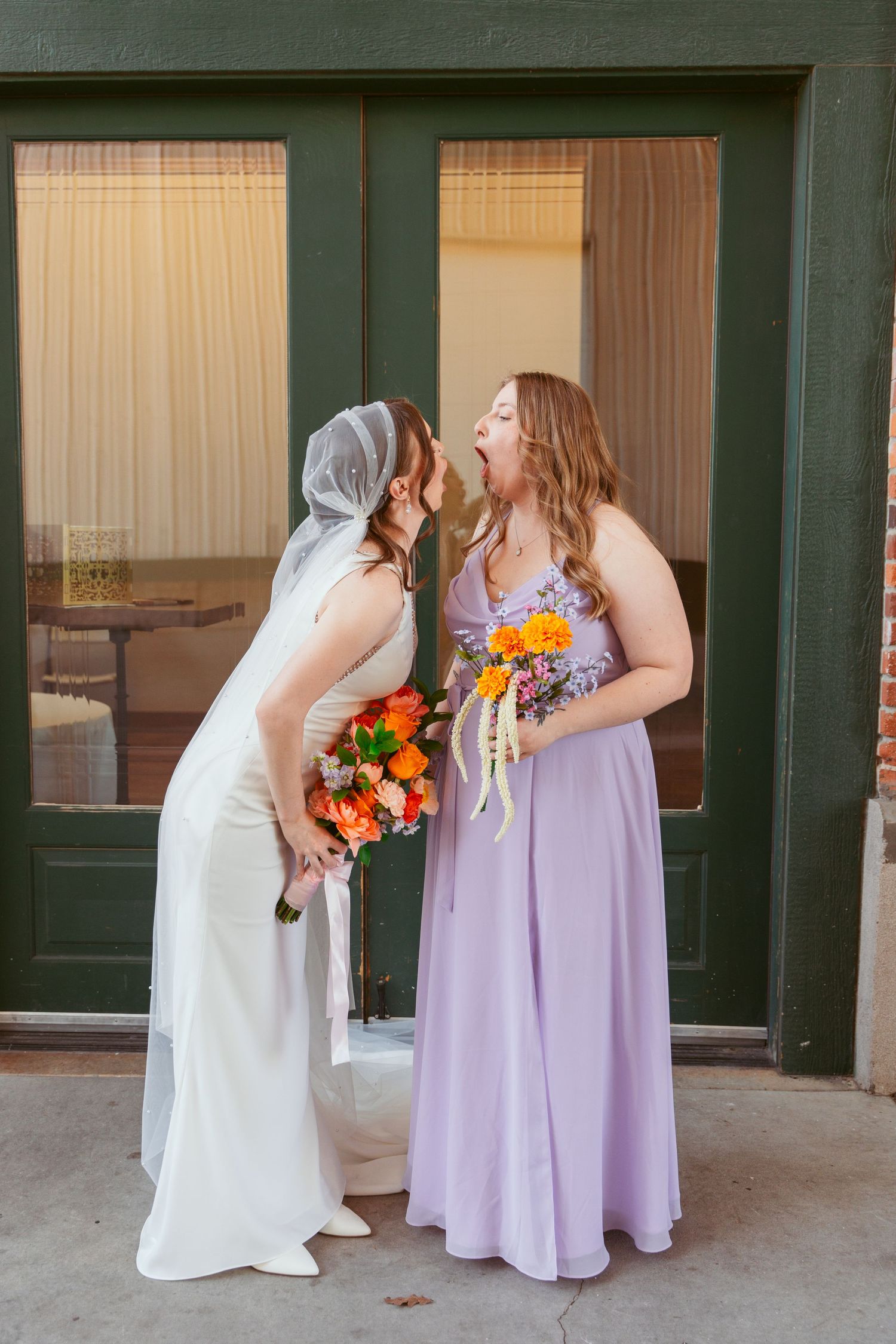Two people in wedding attire share laughter against green door backdrop.