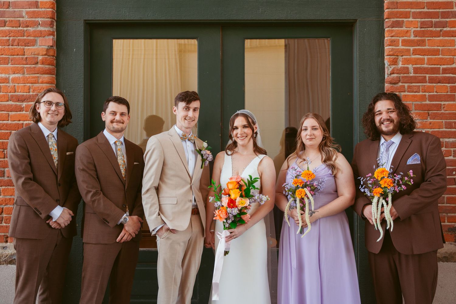 Wedding group photo with bridal party in purple dresses and brown suits against brick building.
