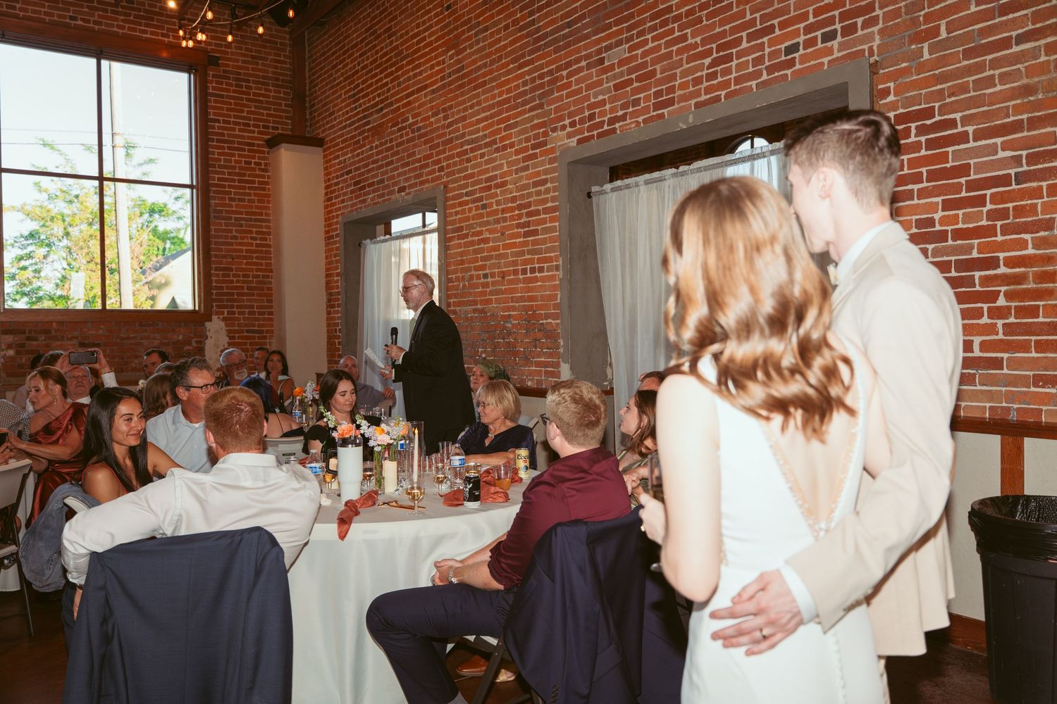 Wedding guests seated at round tables in an industrial brick venue with tall windows and string lights.
