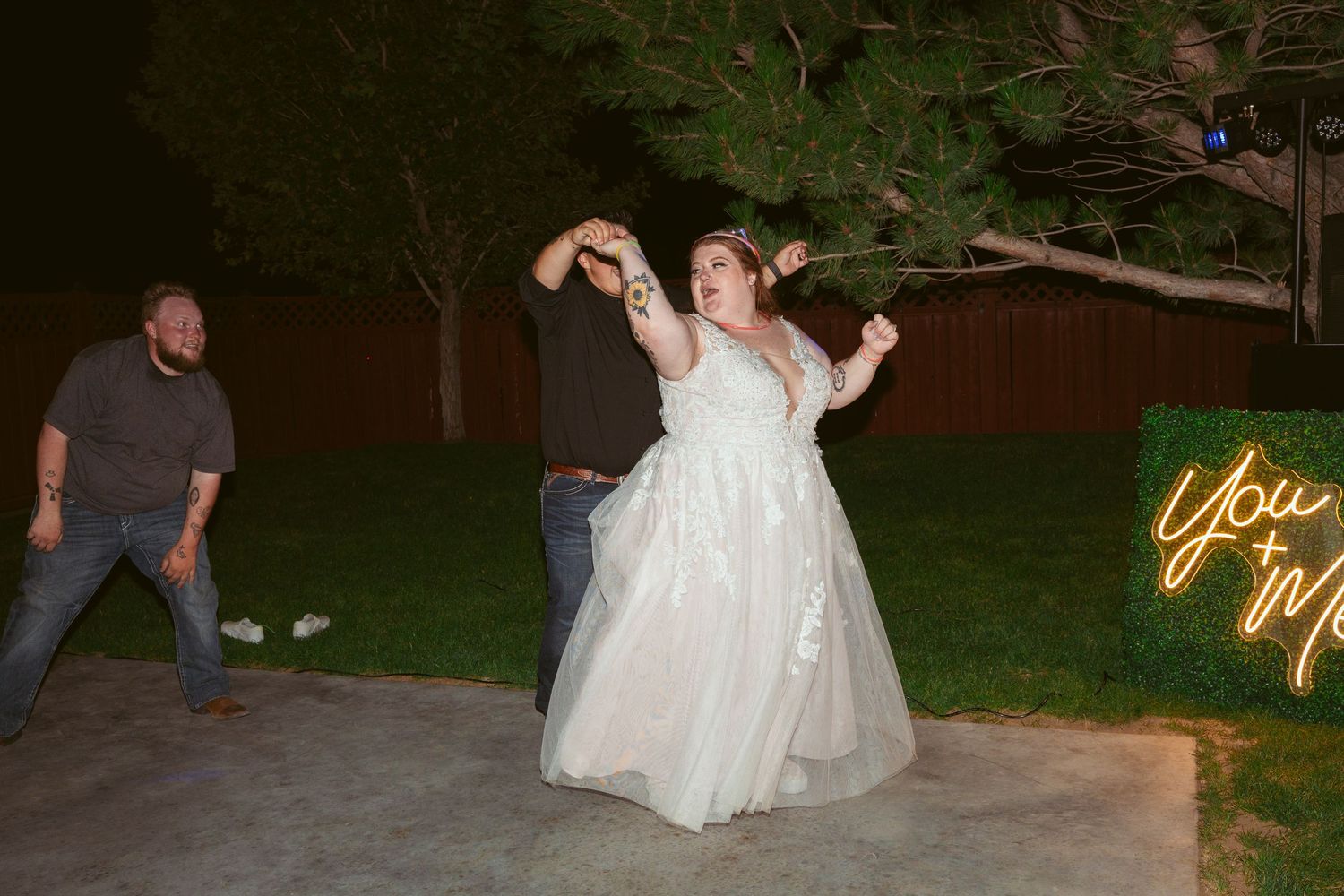 A person in a white wedding dress dances energetically on a patio at night with a neon sign glowing in the background.