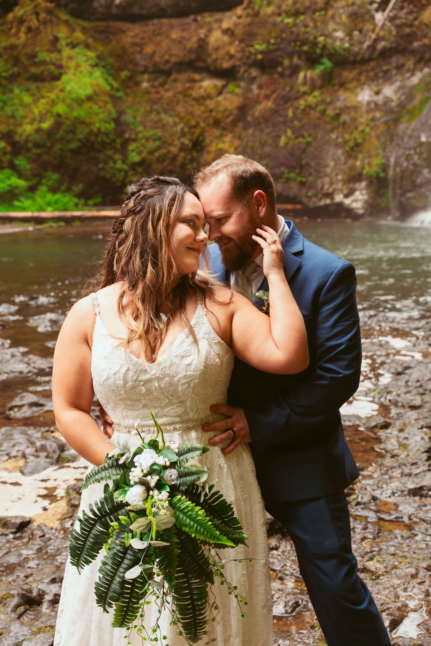 A tender moment between newlyweds by a waterfall with a fern bouquet.
