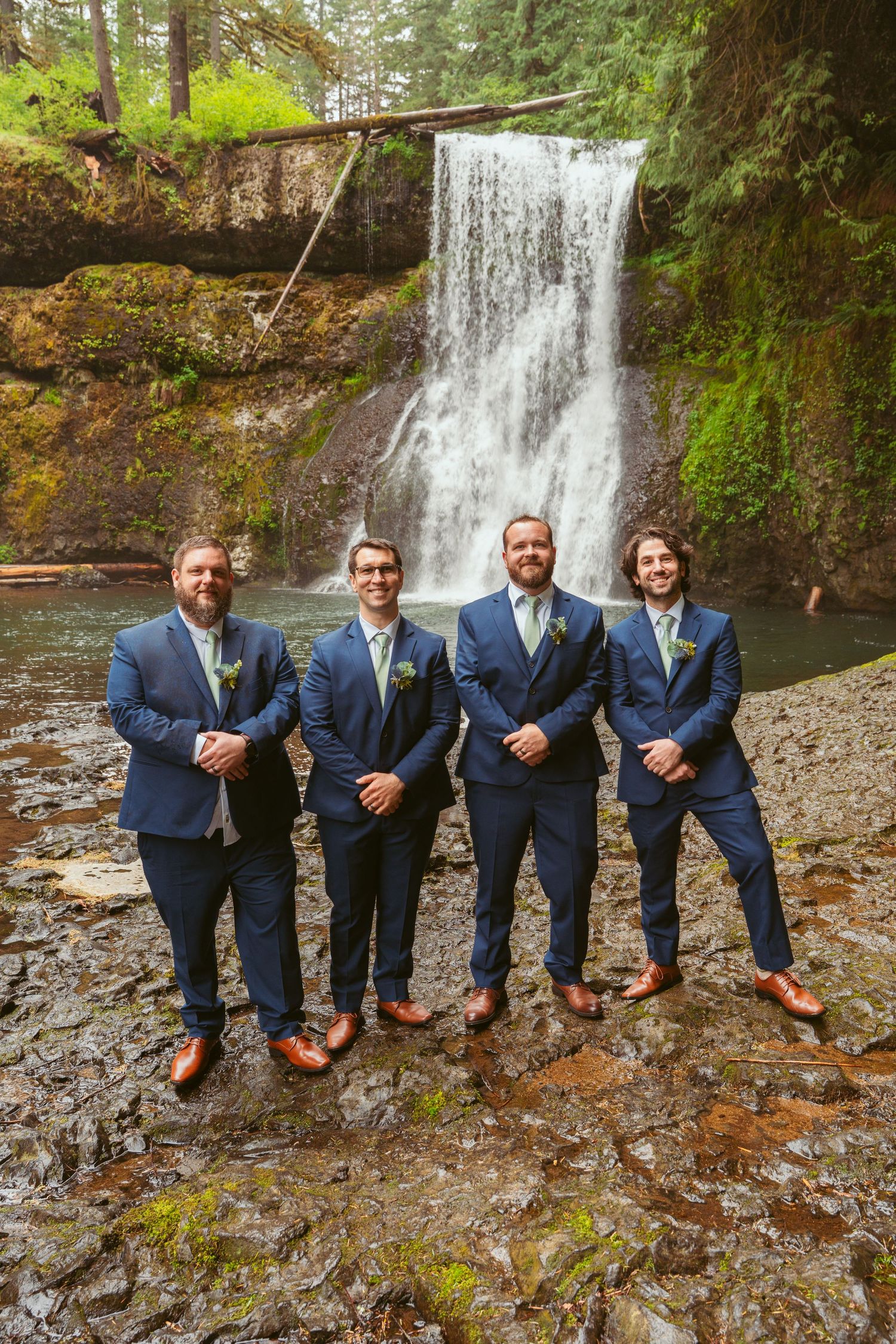 Groomsmen sit casually on rocks near a waterfall wearing coordinated navy suits.