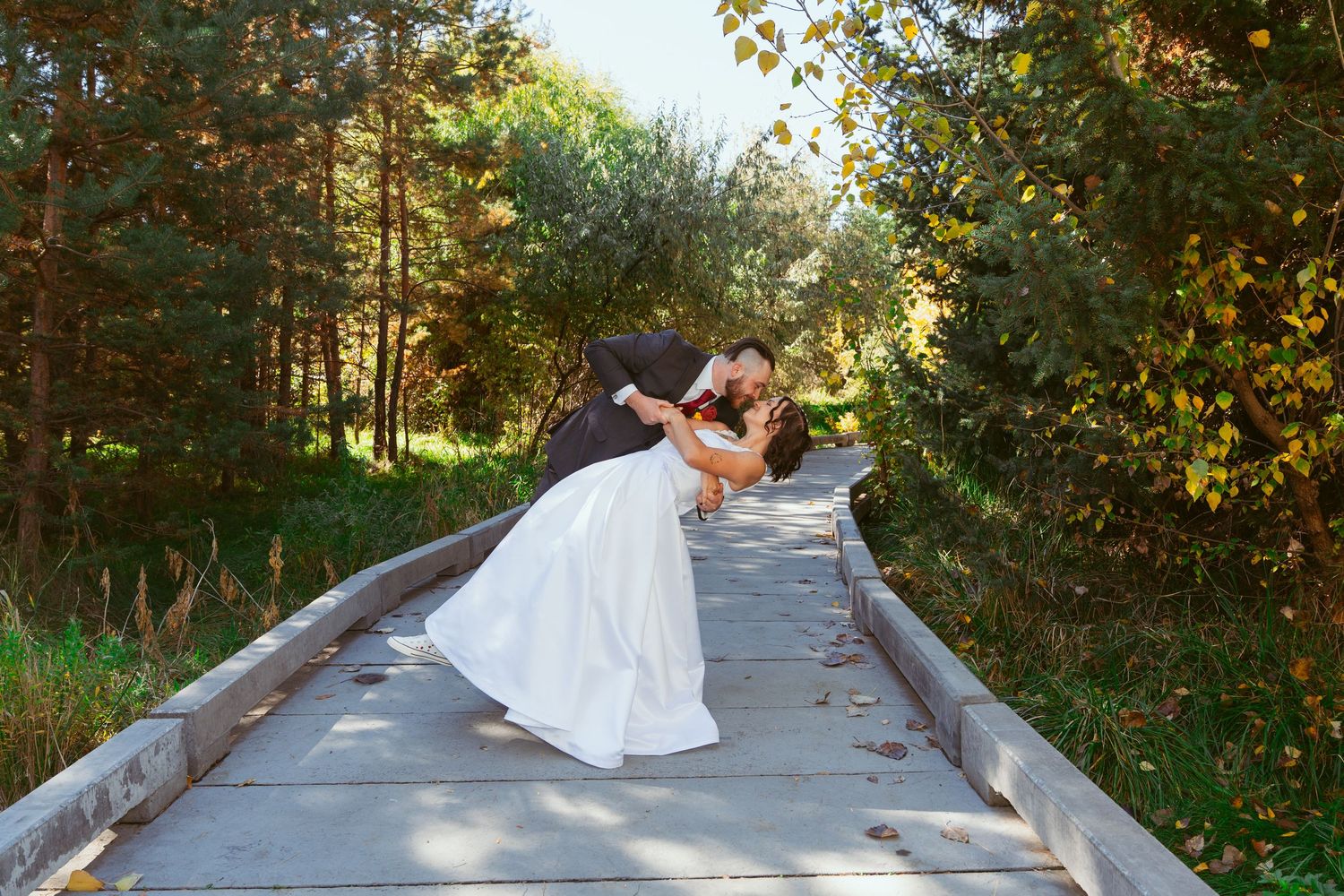 Newlyweds share a romantic dip pose on a wooden walkway surrounded by trees.